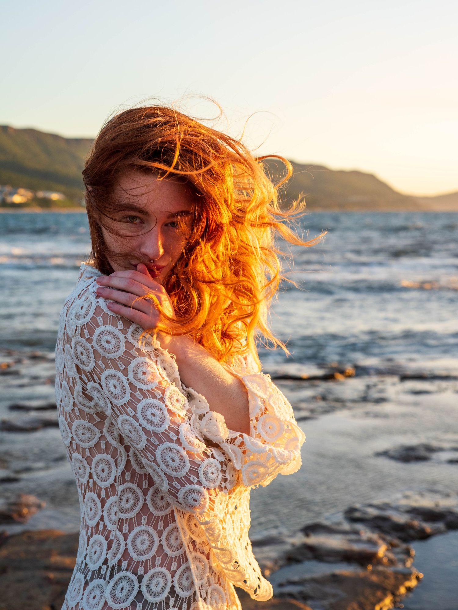 Coastal photoshoot featuring white lace dress and windswept hair at golden hour.