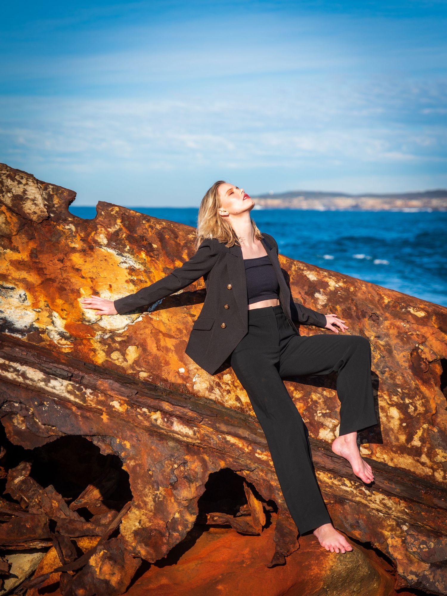 Fashion shoot on coastal rocks with model in black suit against blue sky and ocean.