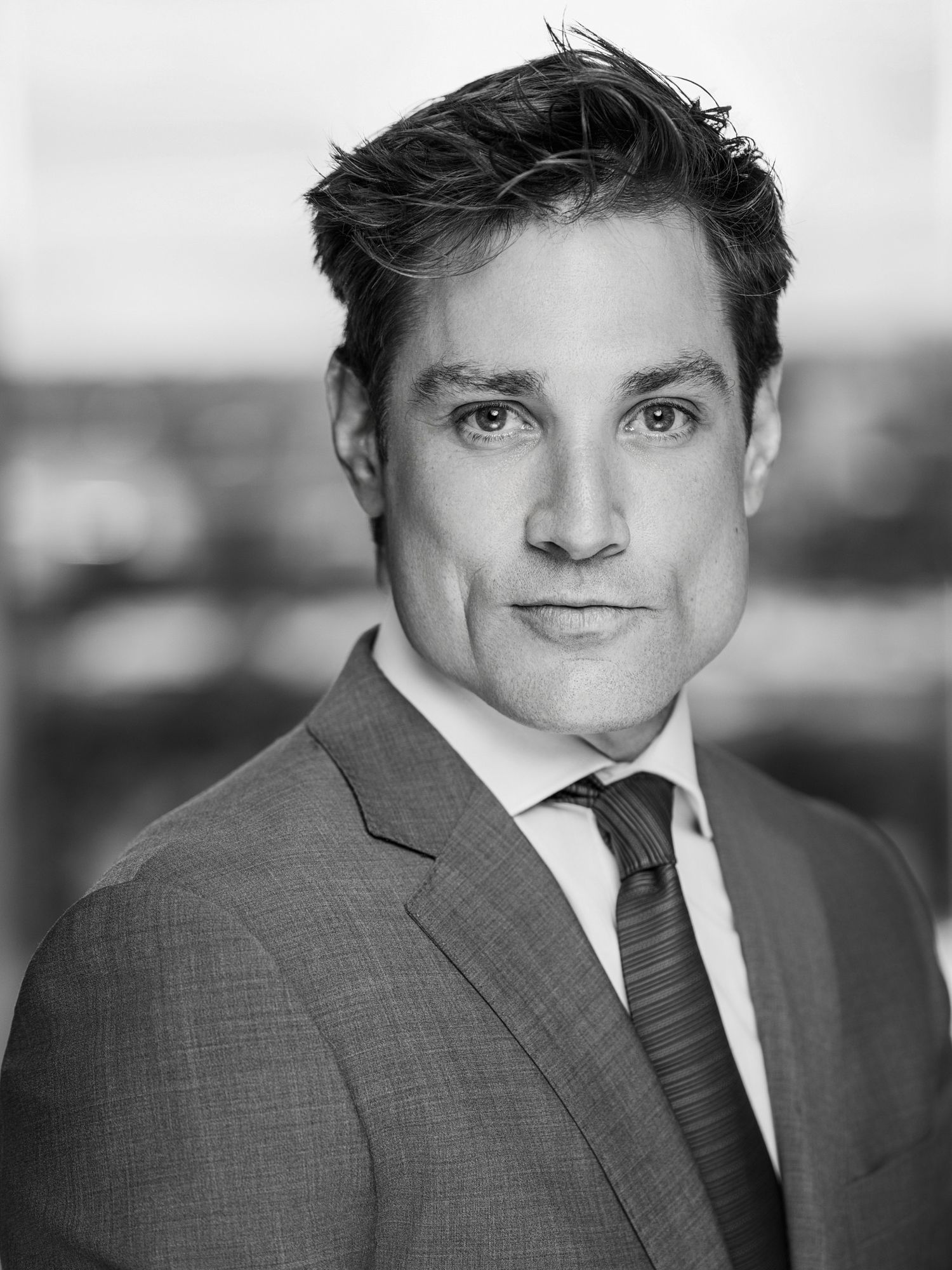 Professional headshot of person in grey suit with dark tie against light background.