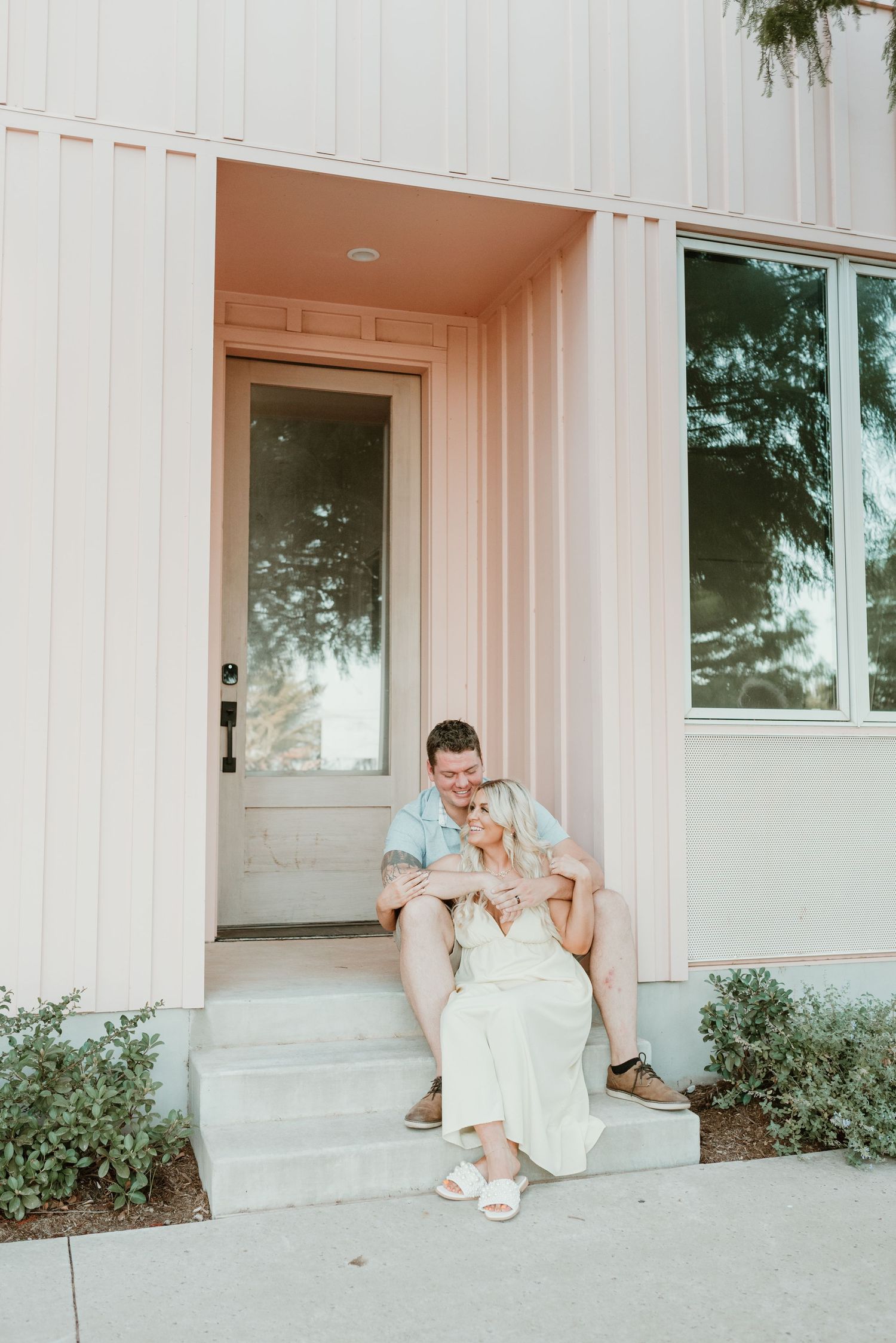 A couple sits together on front steps of a modern pink building, smiling and embracing.