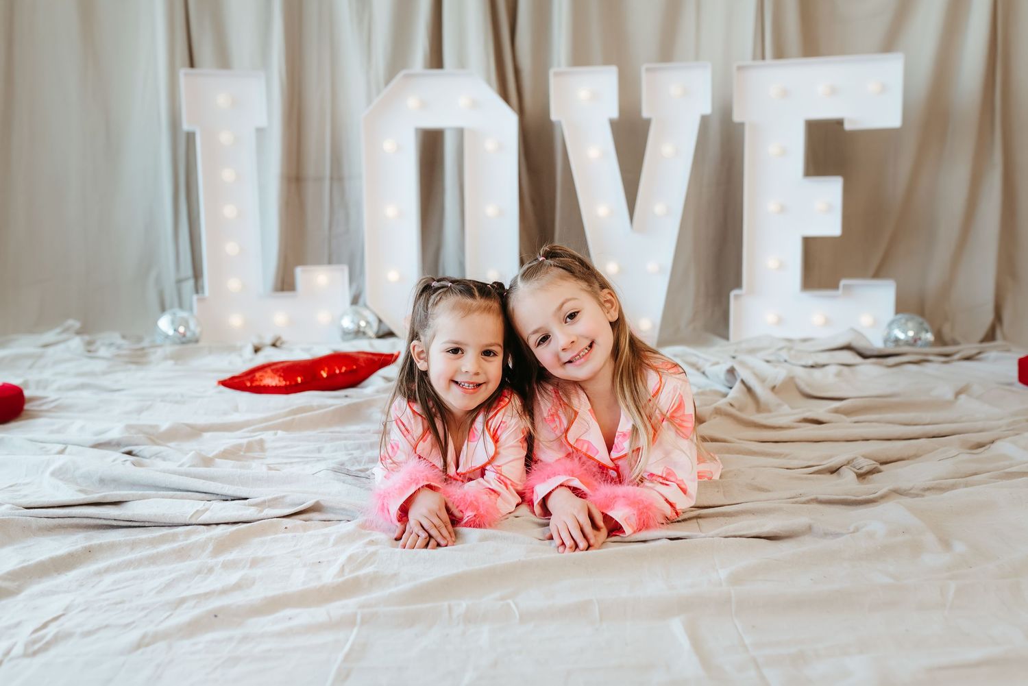 Two young girls in matching pink pajamas pose lying down before a lit LOVE sign backdrop.
