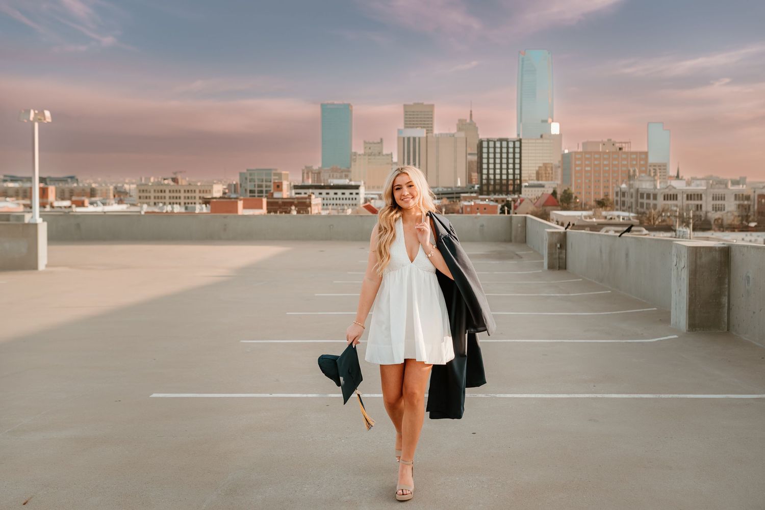 Blonde woman in white dress holding graduation cap and gown on rooftop with city skyline at sunset.