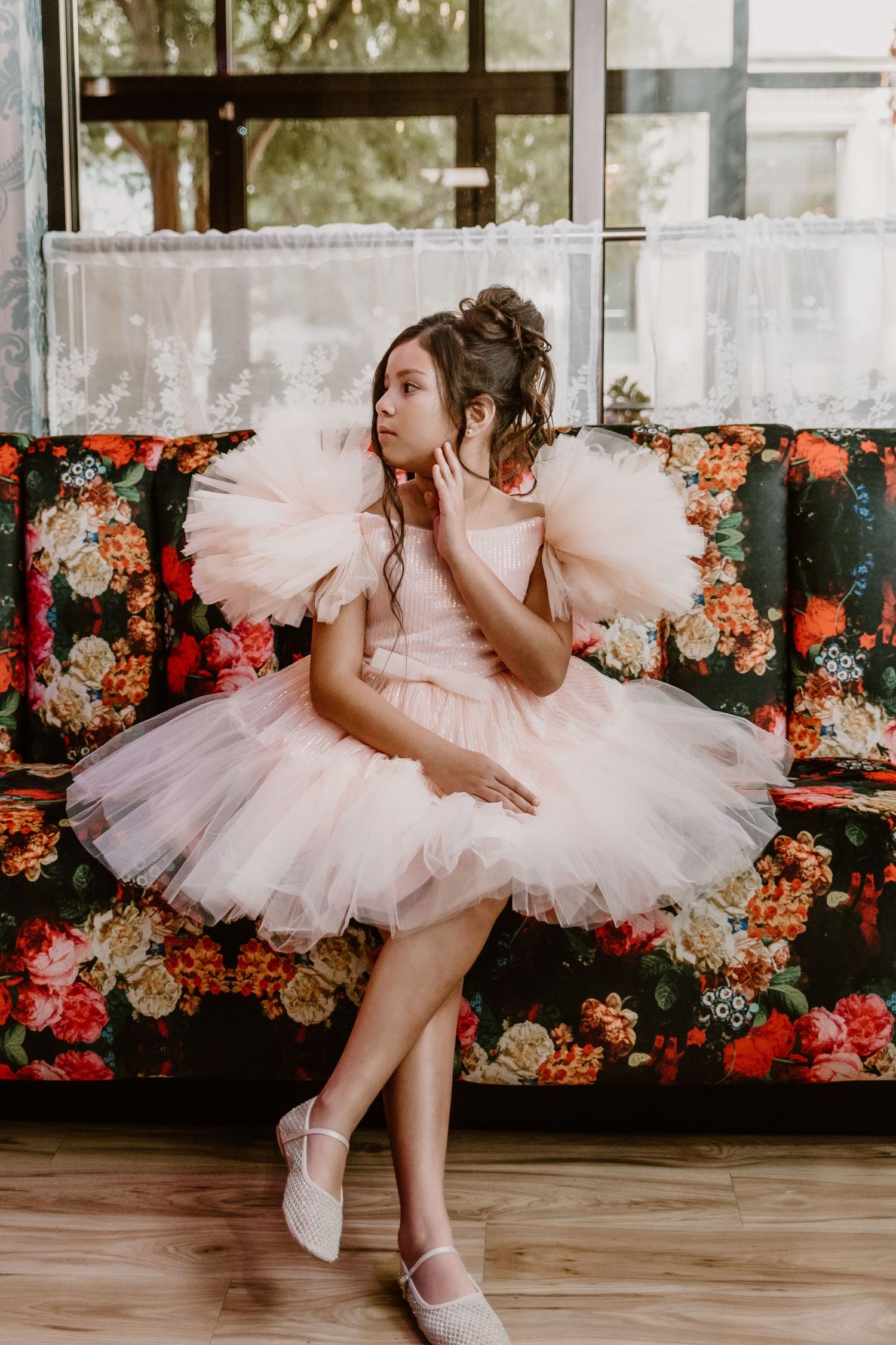 Young girl in pink tutu dress sitting on floral couch near window in elegant portrait setting.