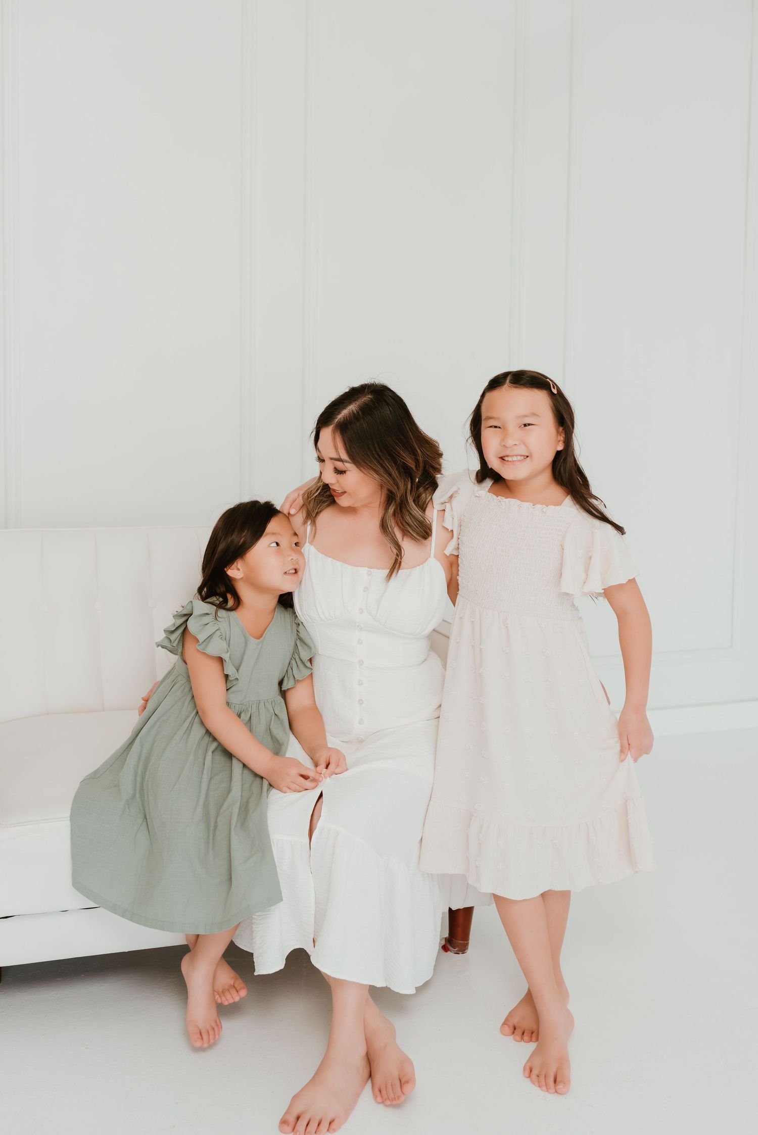 A mother in white dress sits with two daughters in sage green and blush pink dresses in a bright white studio.
