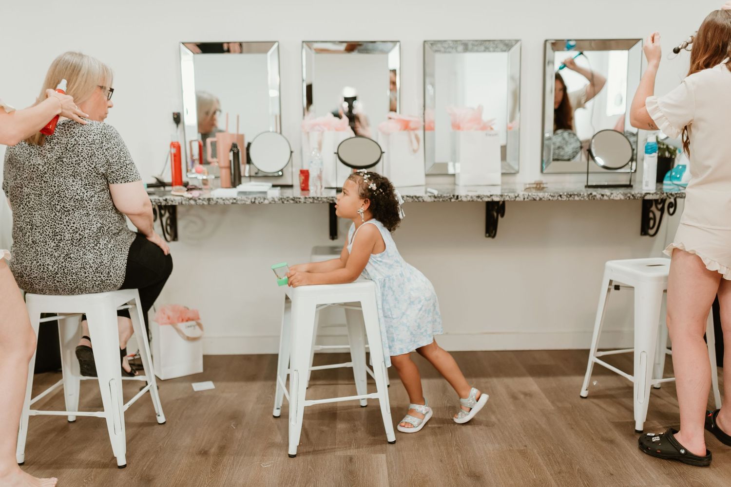Young flower girl sitting at bridal salon vanity mirror while stylists prepare for wedding day.