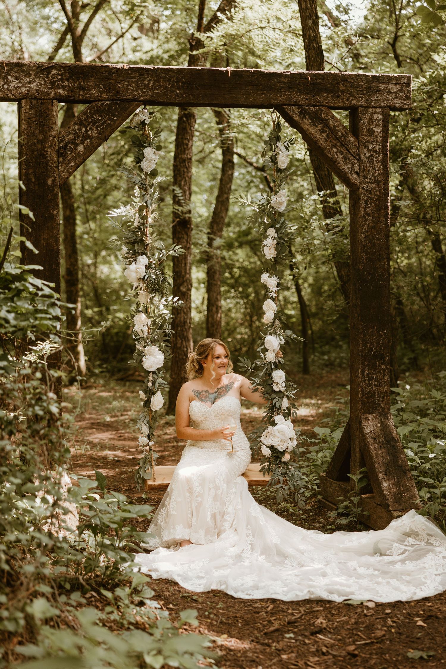 Bride in lace gown sitting on wooden swing adorned with white roses in a lush forest setting.