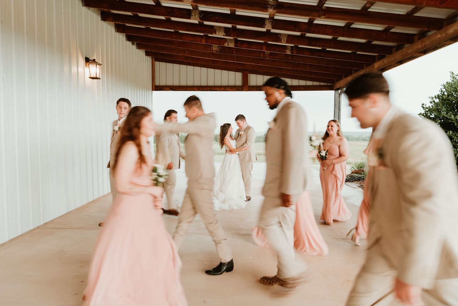 Wedding party walking under a covered outdoor venue with bridesmaids in blush pink dresses and groomsmen in tan suits.