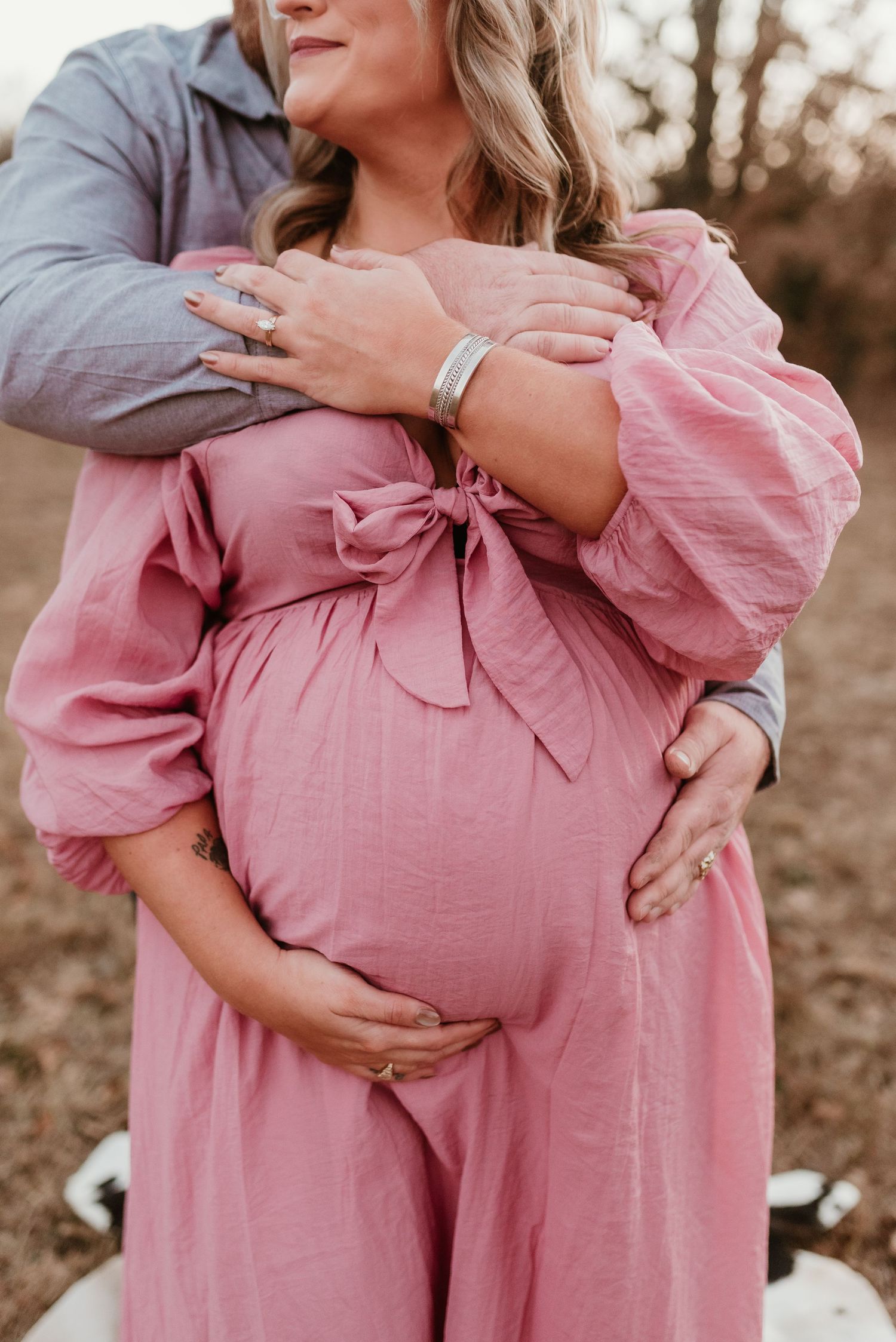 A close-up of a pregnant woman in a pink dress cradling her baby bump while her partner embraces her from behind.