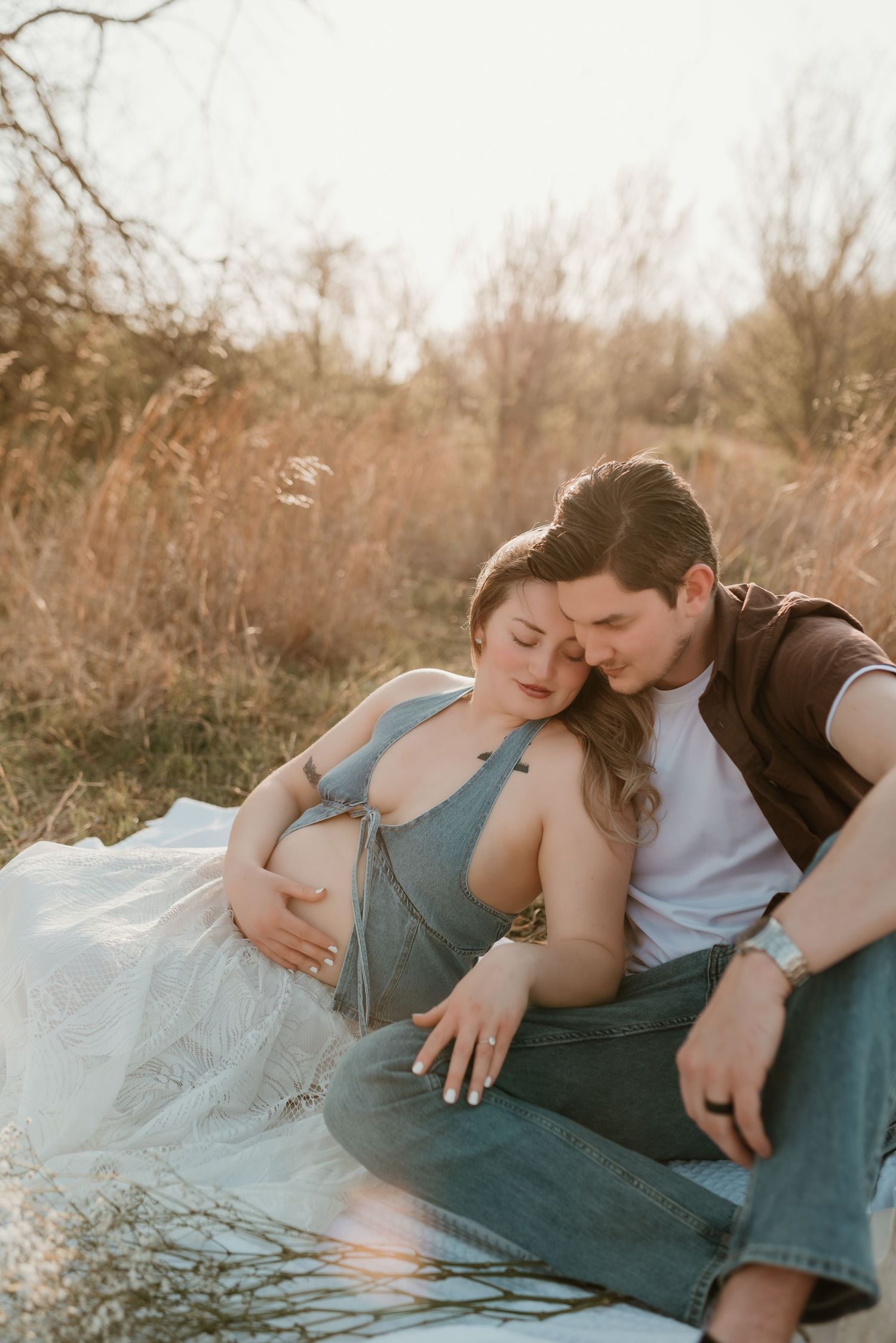 Couple sitting outdoors in golden light, man embracing pregnant woman in blue dress among tall grass.