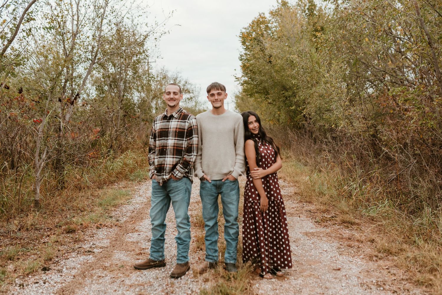 Three people posing on a rustic dirt path surrounded by autumn foliage for a fall family photo session.