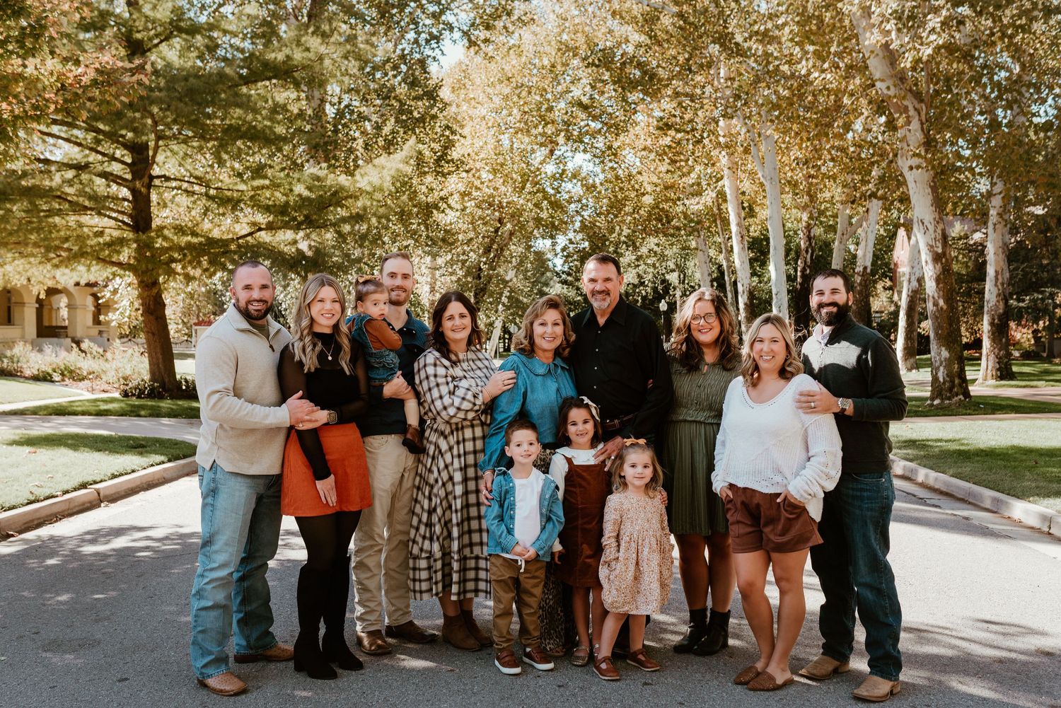 Extended family of twelve smiling together outdoors surrounded by vibrant fall foliage.