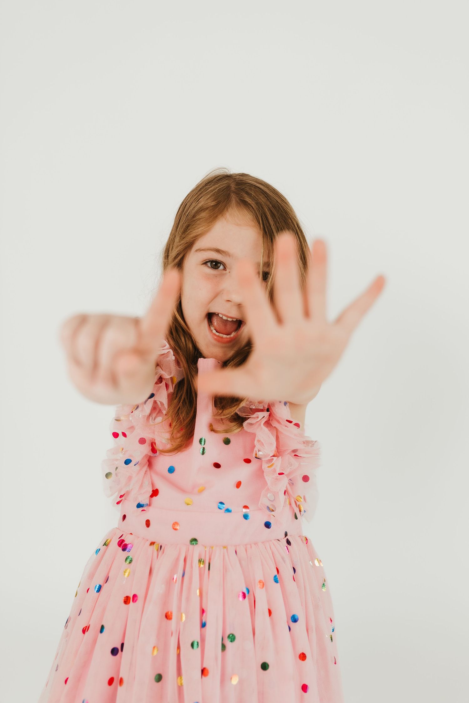 A young girl in a pink confetti dress reaches her hand toward the camera with an excited expression.