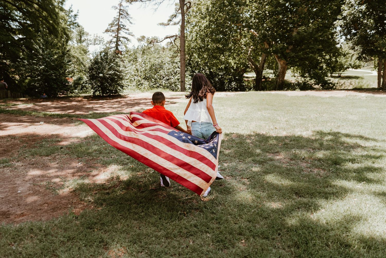 Two children run through a park holding a large American flag on a sunny day.