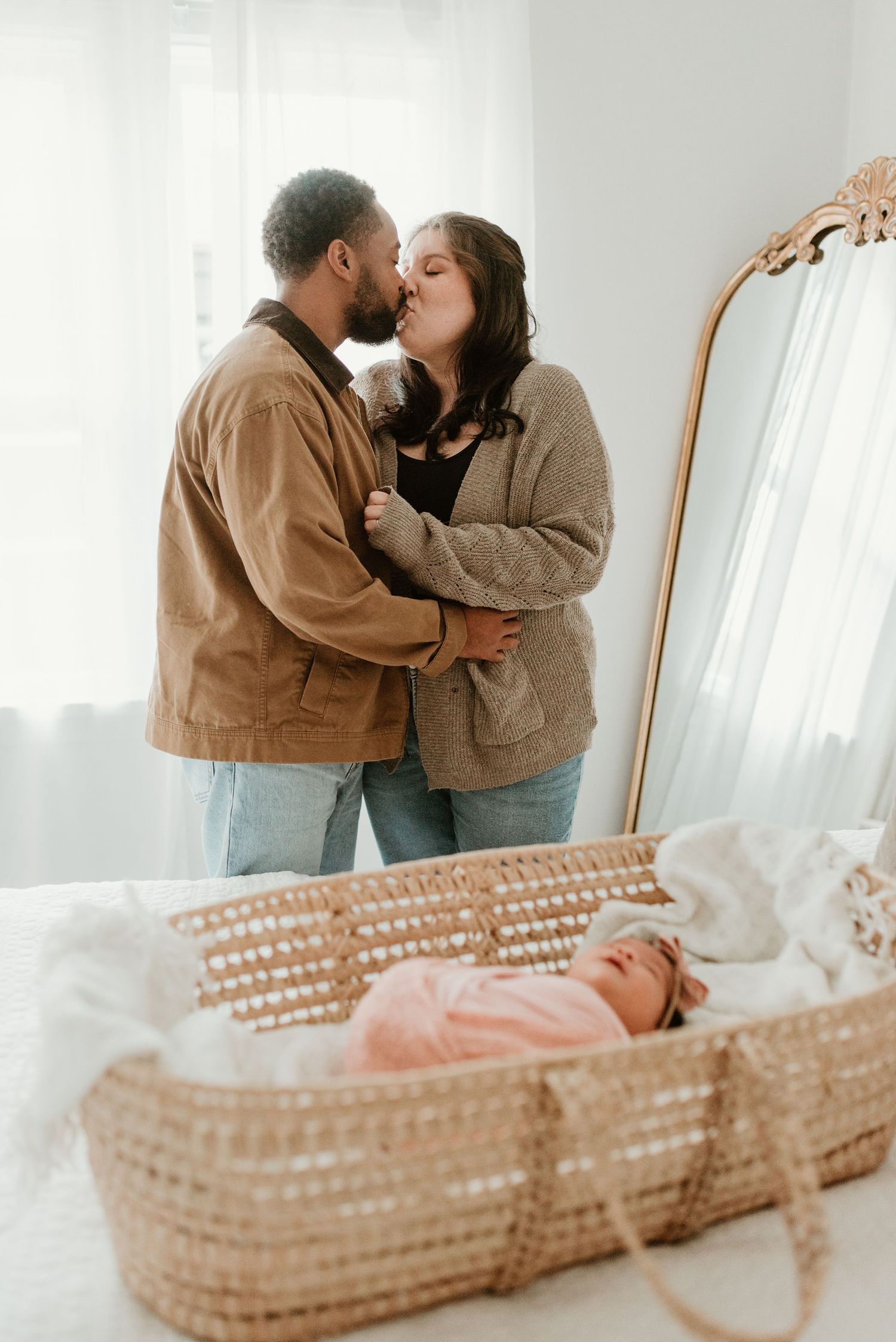Couple sharing a kiss while their newborn baby sleeps peacefully in a wicker moses basket.