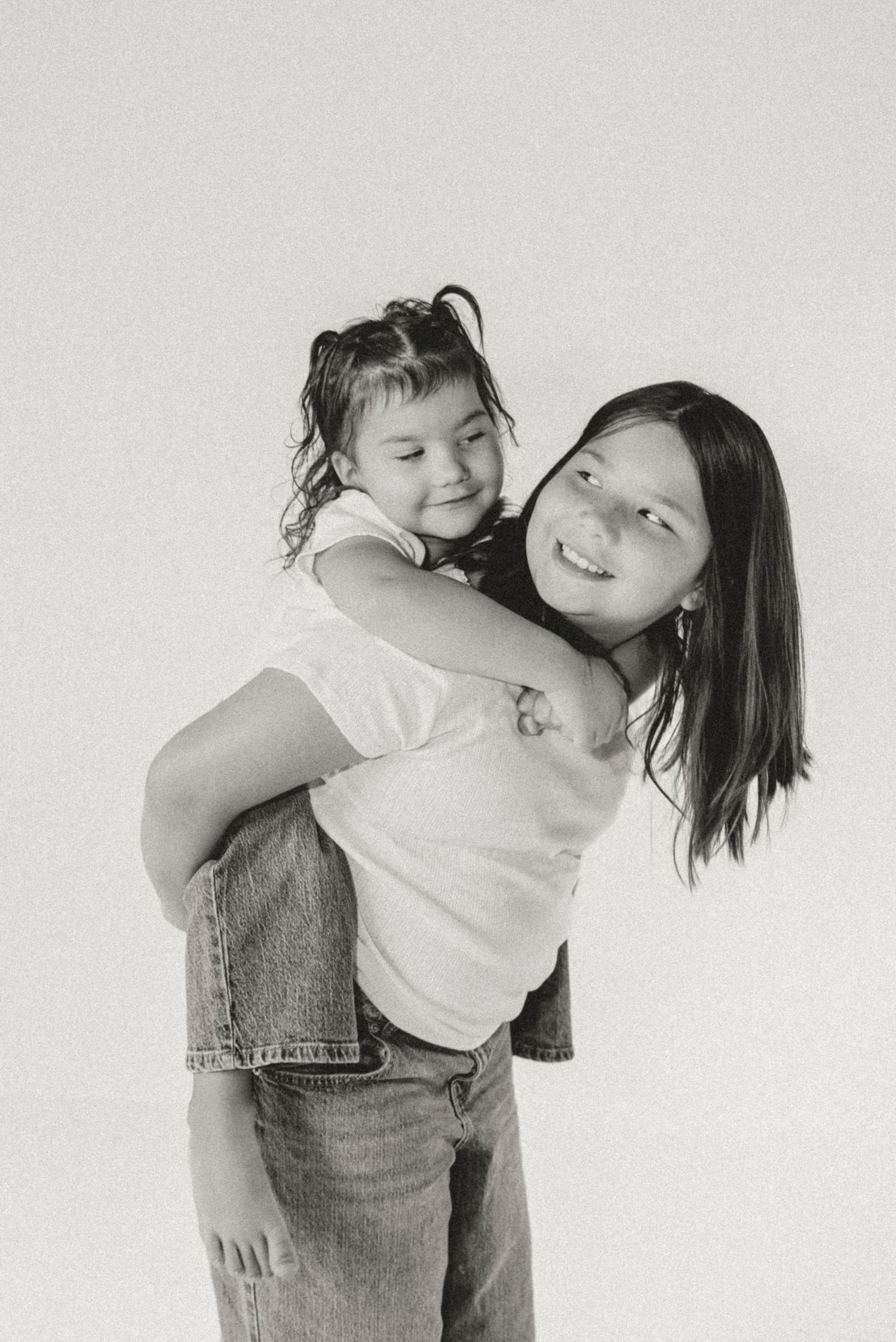 Black and white photo of older sister giving younger sibling a piggyback ride, both smiling happily.