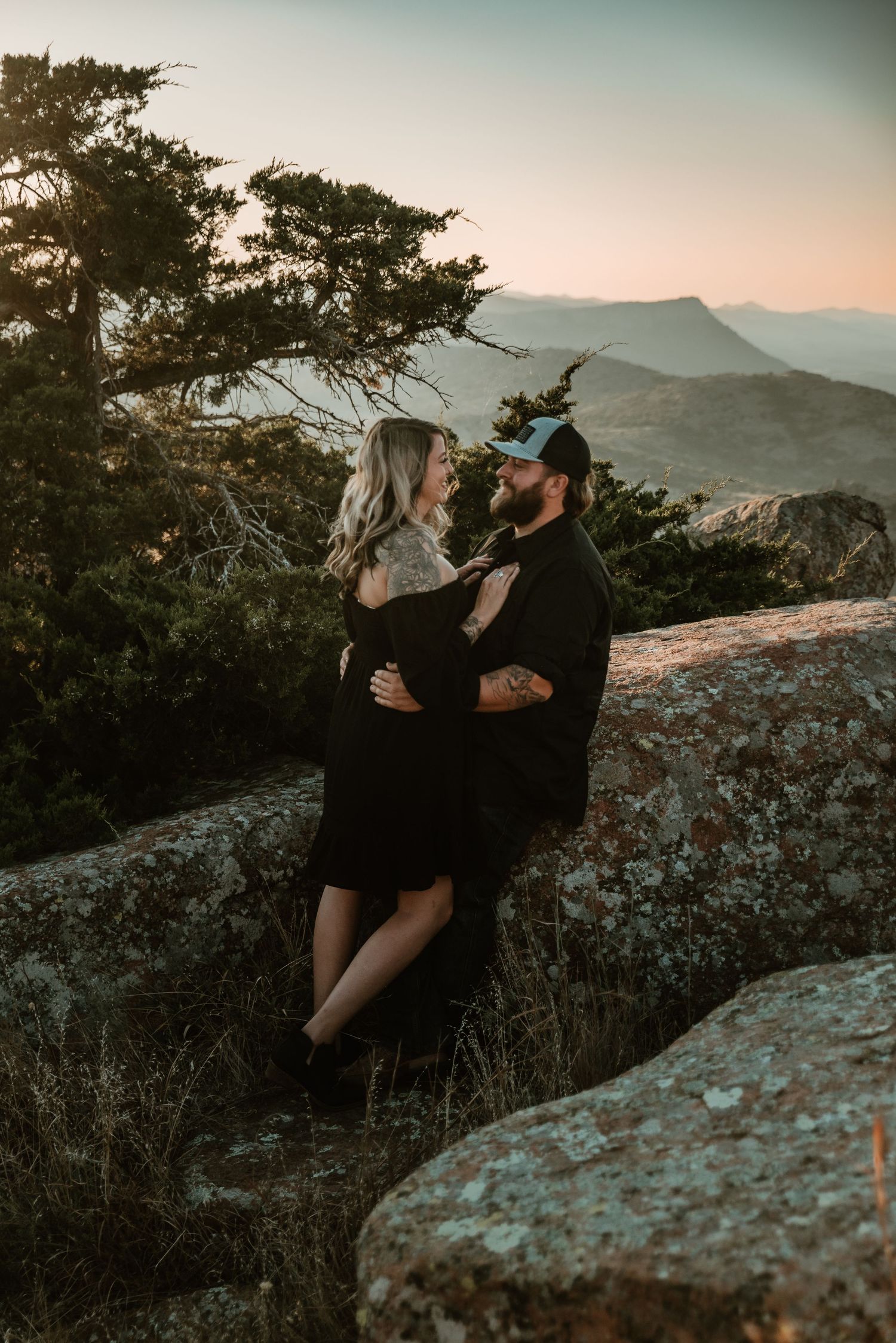 Couple embracing on rocky mountain overlook at sunset, woman in black dress, man in black shirt and blue cap.