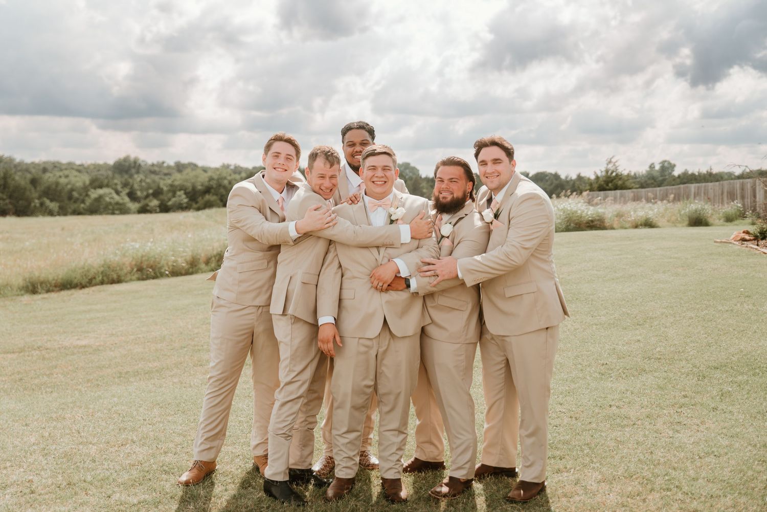 Groom and groomsmen in matching tan suits posing playfully together on a grassy outdoor field.