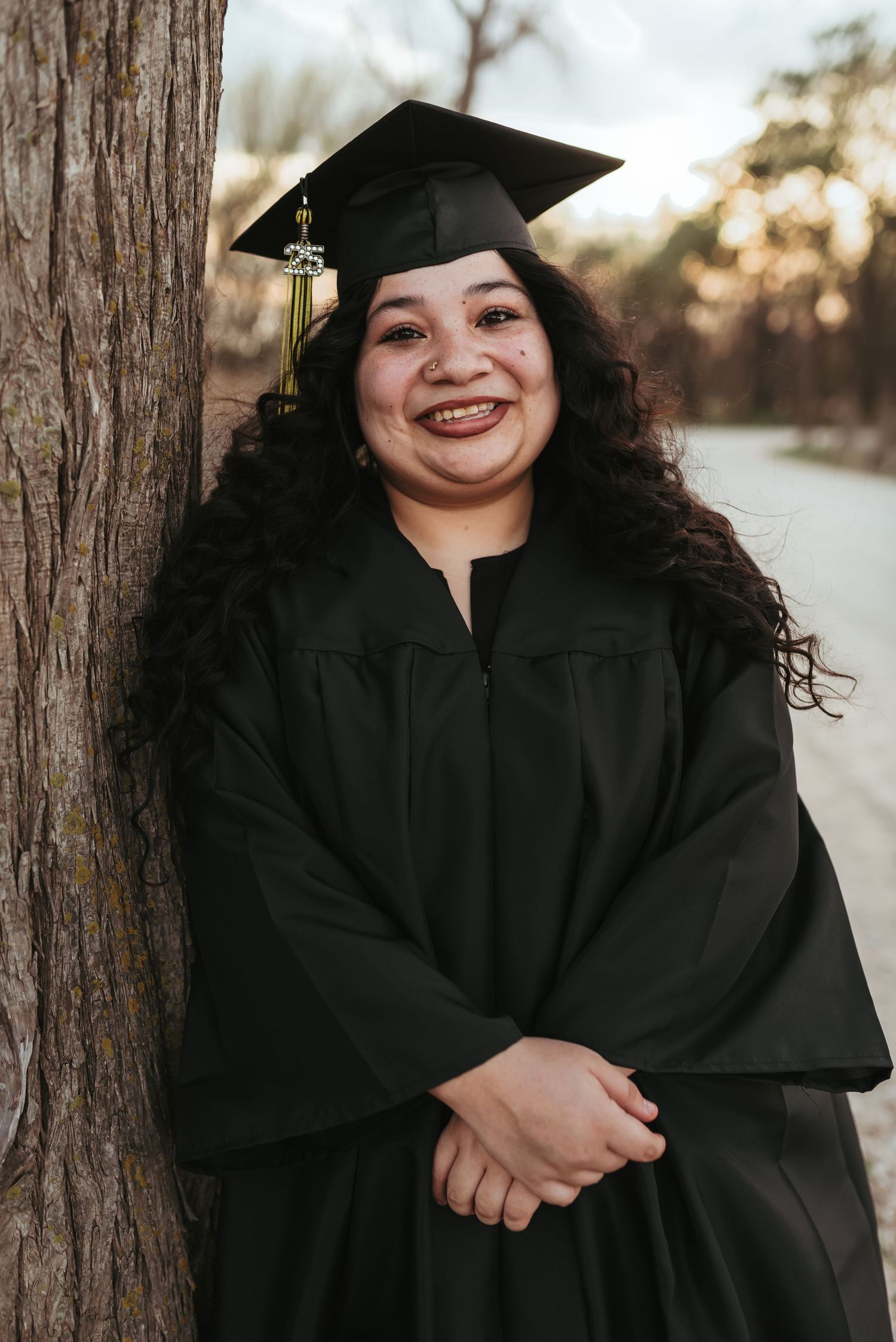 A smiling graduate in black cap and gown poses outdoors leaning against a tree.