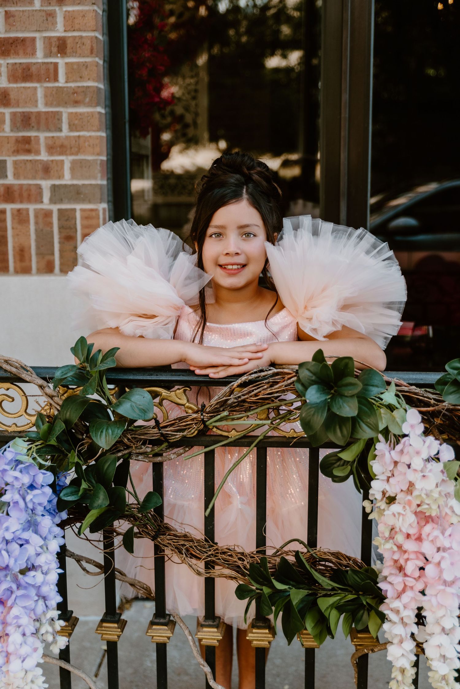 Smiling young girl in pink tutu dress leaning on iron railing decorated with colorful flowers.