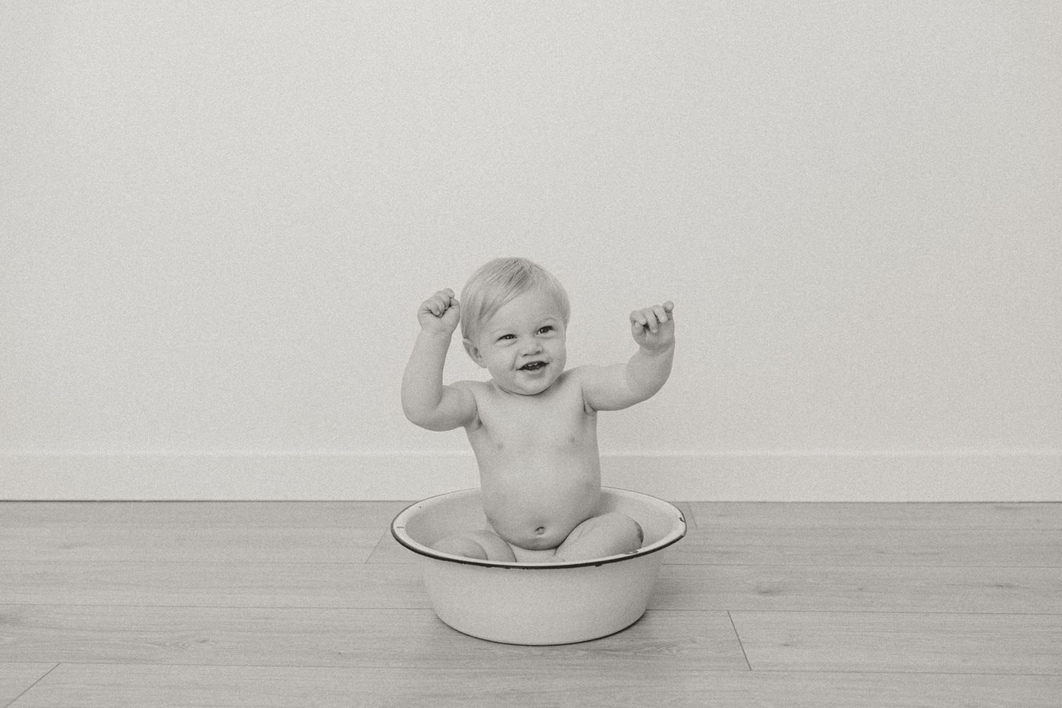 Black and white photo of joyful baby sitting in enamel tub with arms raised in celebration.
