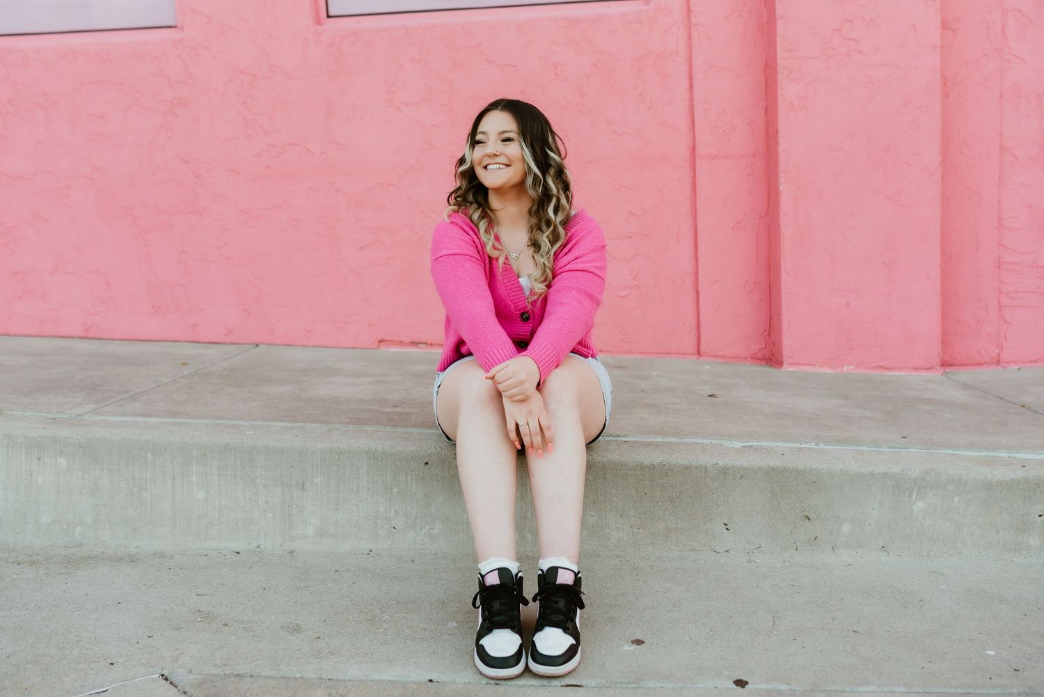 Young woman in pink outfit sitting on concrete steps smiling in front of pink wall.