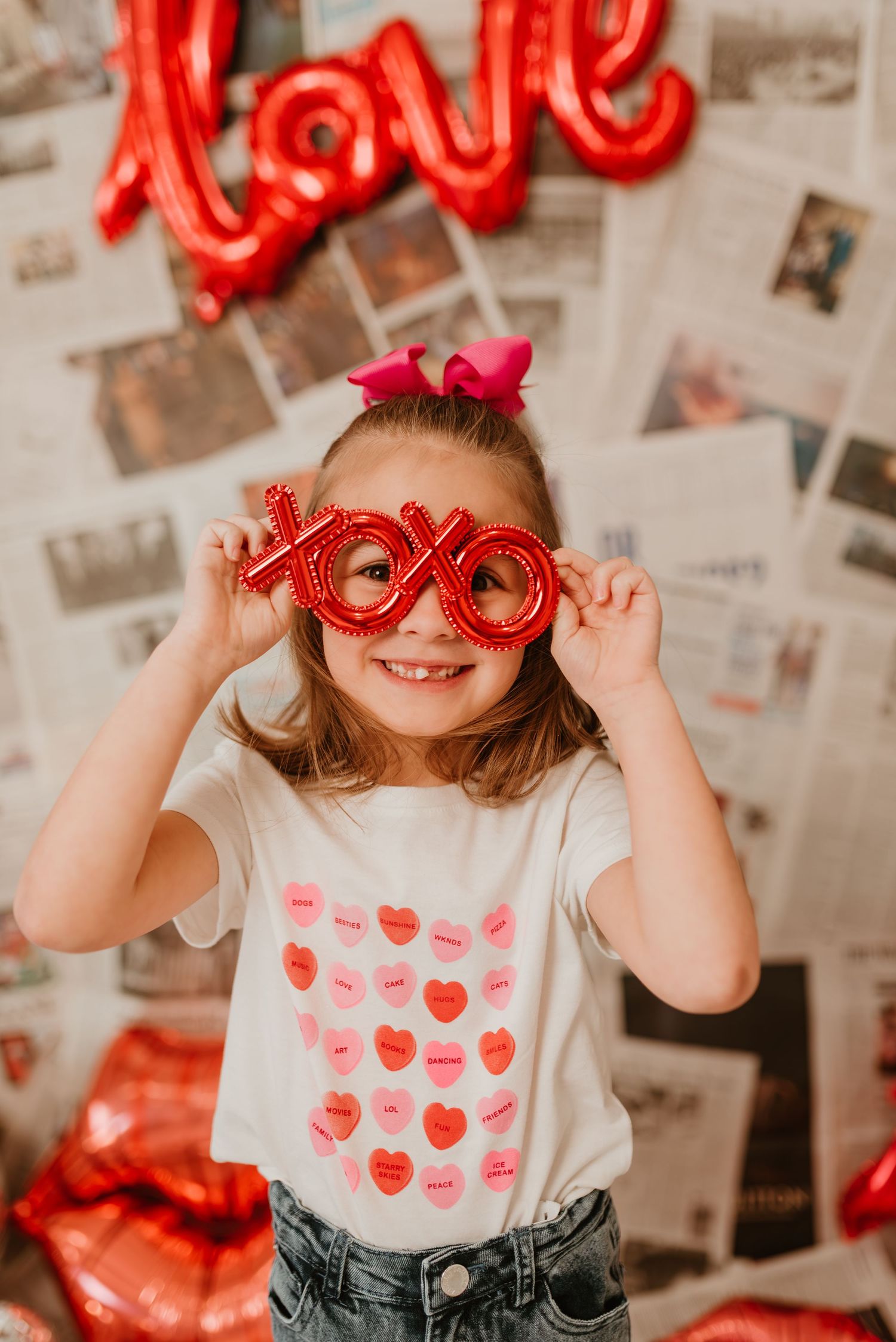 A cheerful girl holds XOXO Valentine glasses wearing a heart-print tee with a love balloon backdrop.
