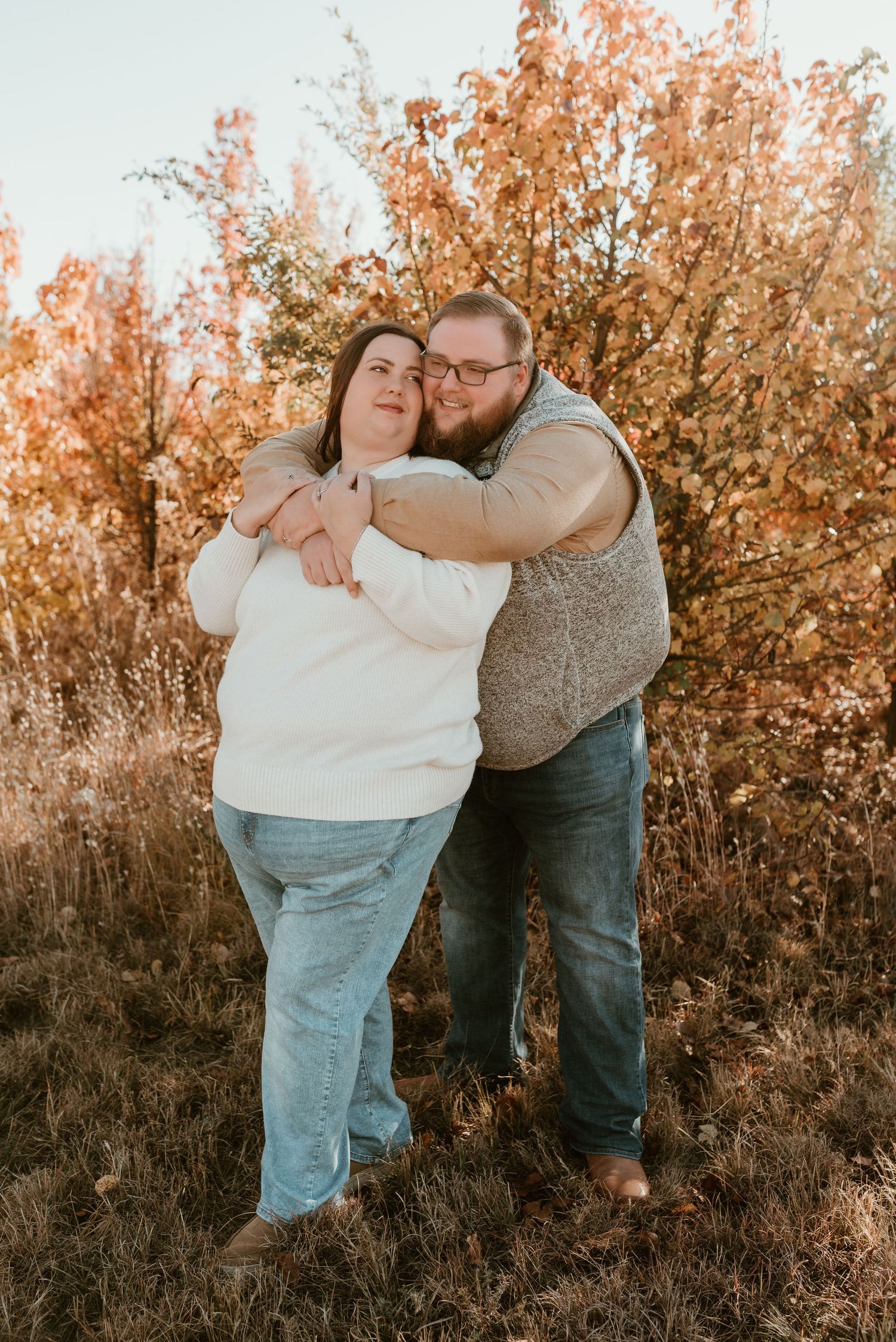 Couple posing together in fall setting, man hugging woman from behind among golden autumn trees.