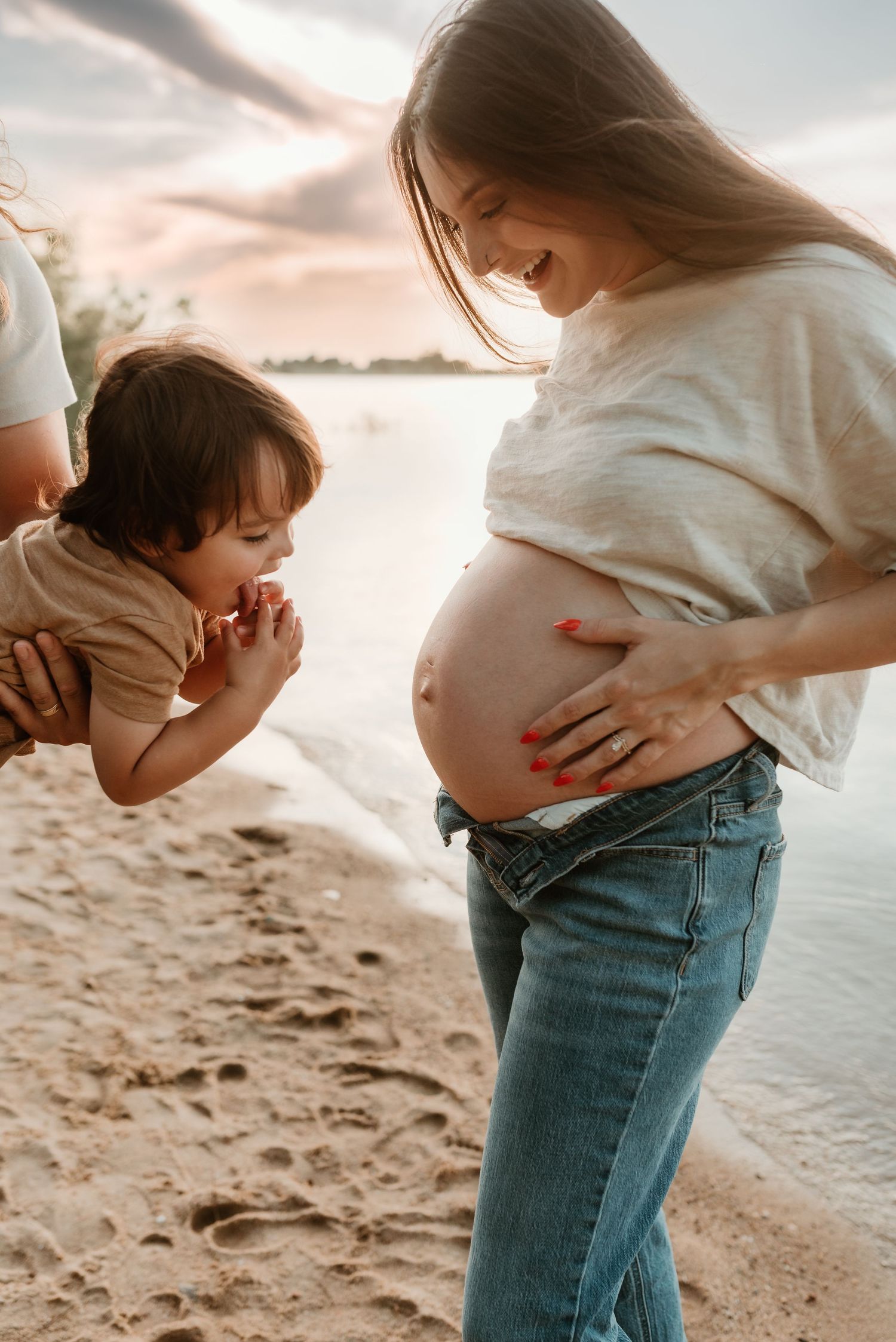 Toddler child curiously examines pregnant mothers bare baby bump on a sandy beach at sunset.
