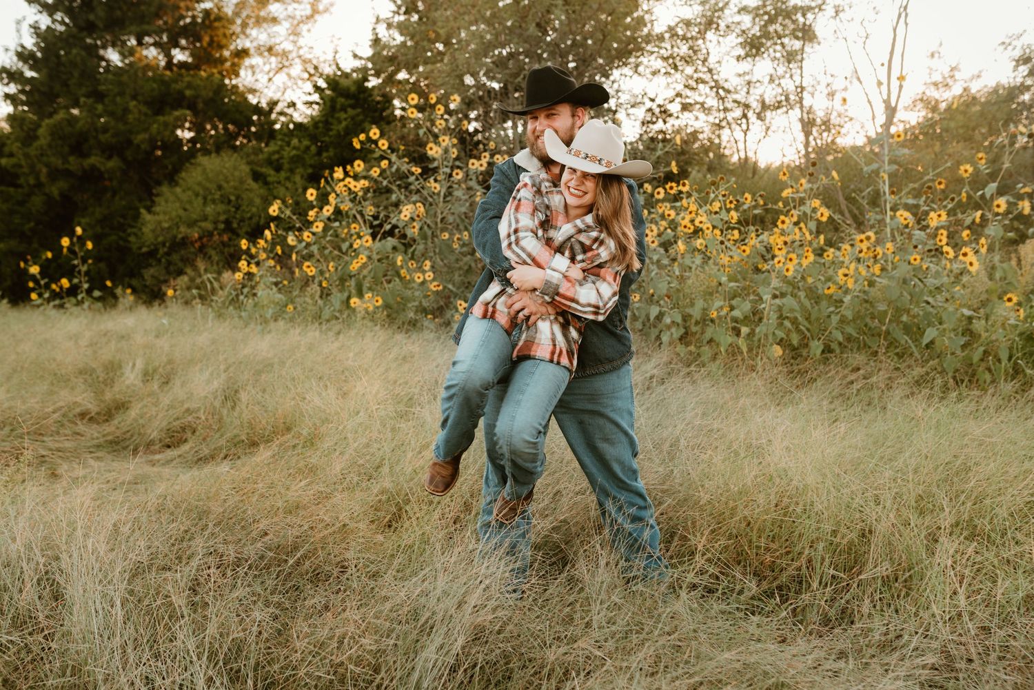 A couple in cowboy hats share a romantic embrace in a golden field with wildflowers.