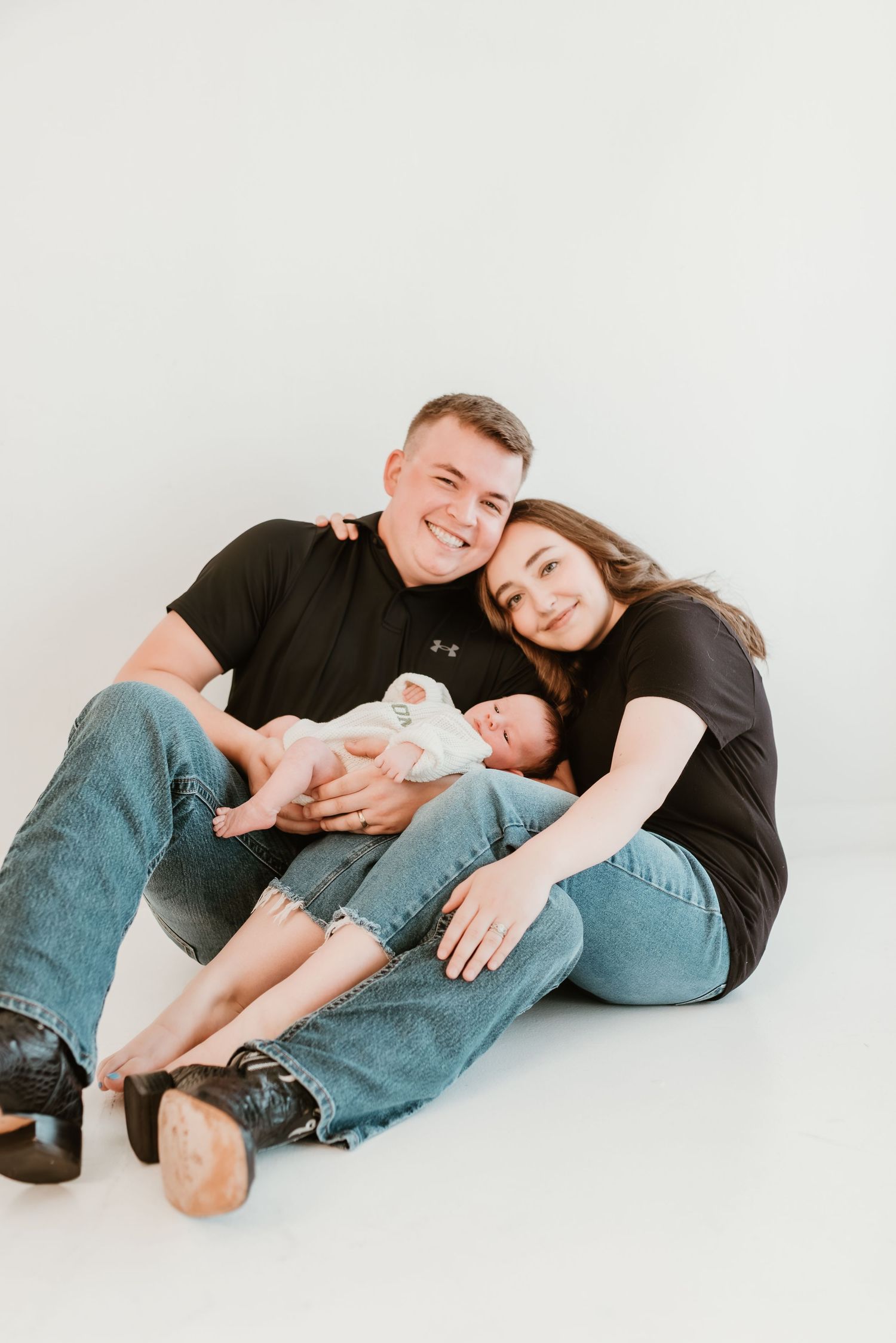 Happy couple sitting on floor in matching black shirts, smiling while cradling their newborn baby.