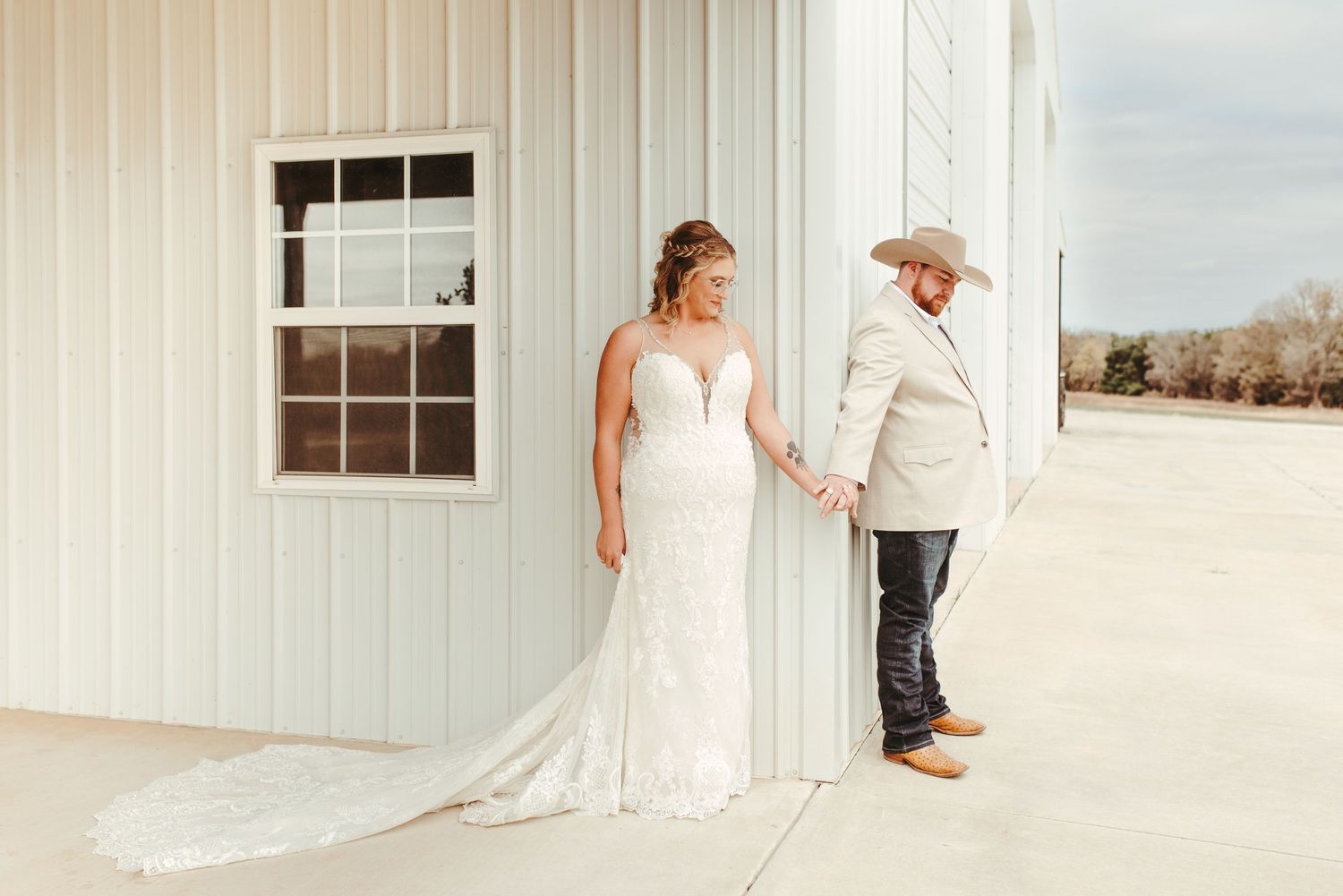 Bride in lace mermaid gown holds hands with groom in tan suit and cowboy hat outside white barn venue.