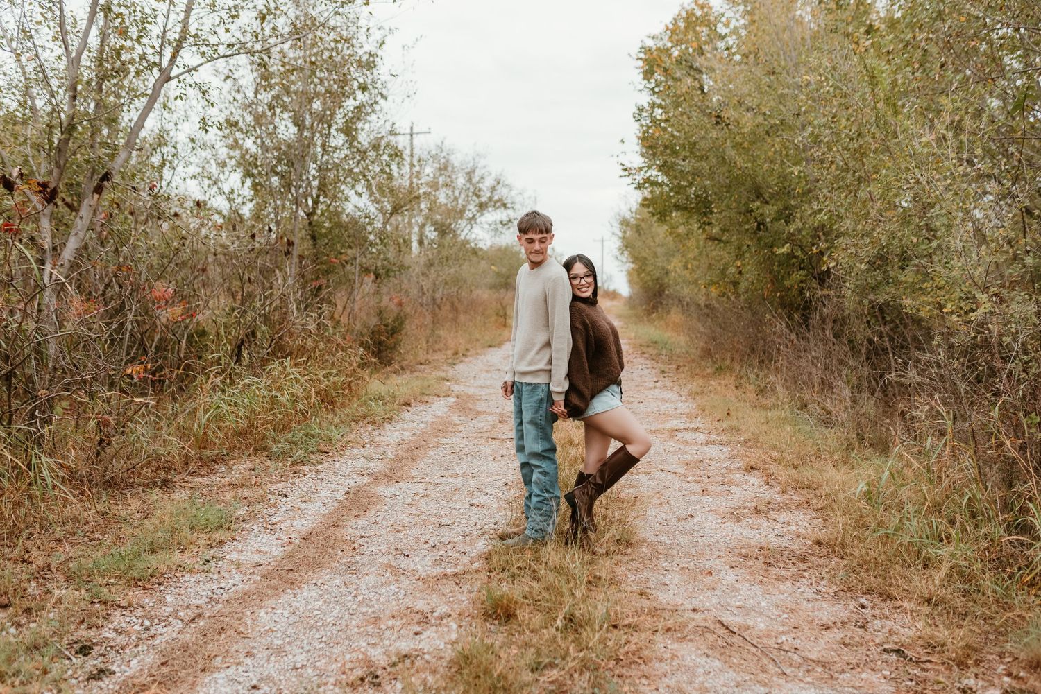 Smiling couple posing together on a scenic dirt trail surrounded by fall trees.