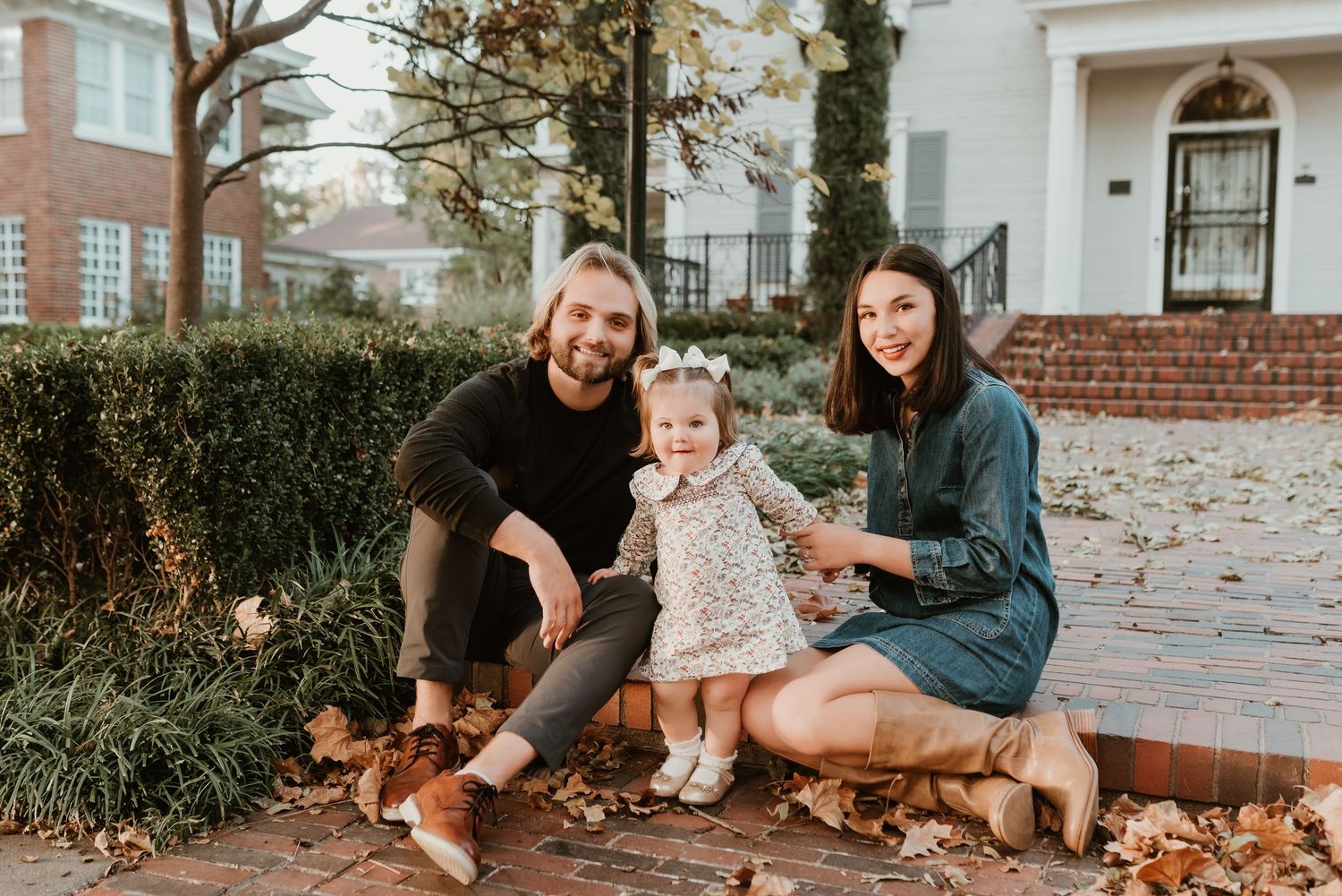 Young family with toddler sits together on brick steps surrounded by autumn leaves.