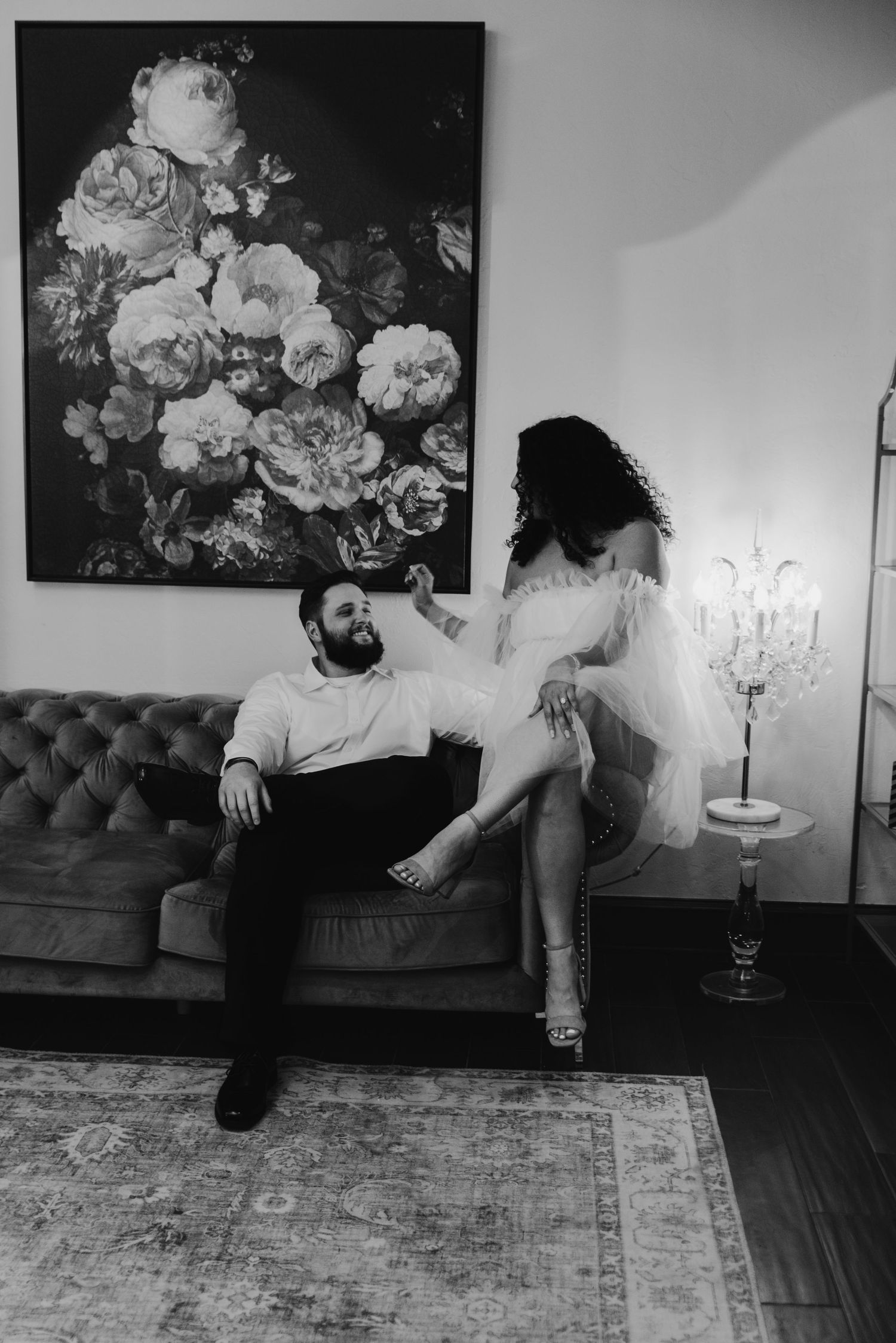 Black and white photo of a couple sharing an intimate moment on a tufted sofa with floral art.