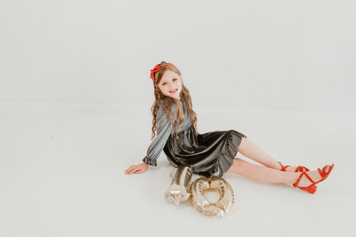 Young girl in a silver dress and red heels sitting on a white studio floor with a gold balloon.
