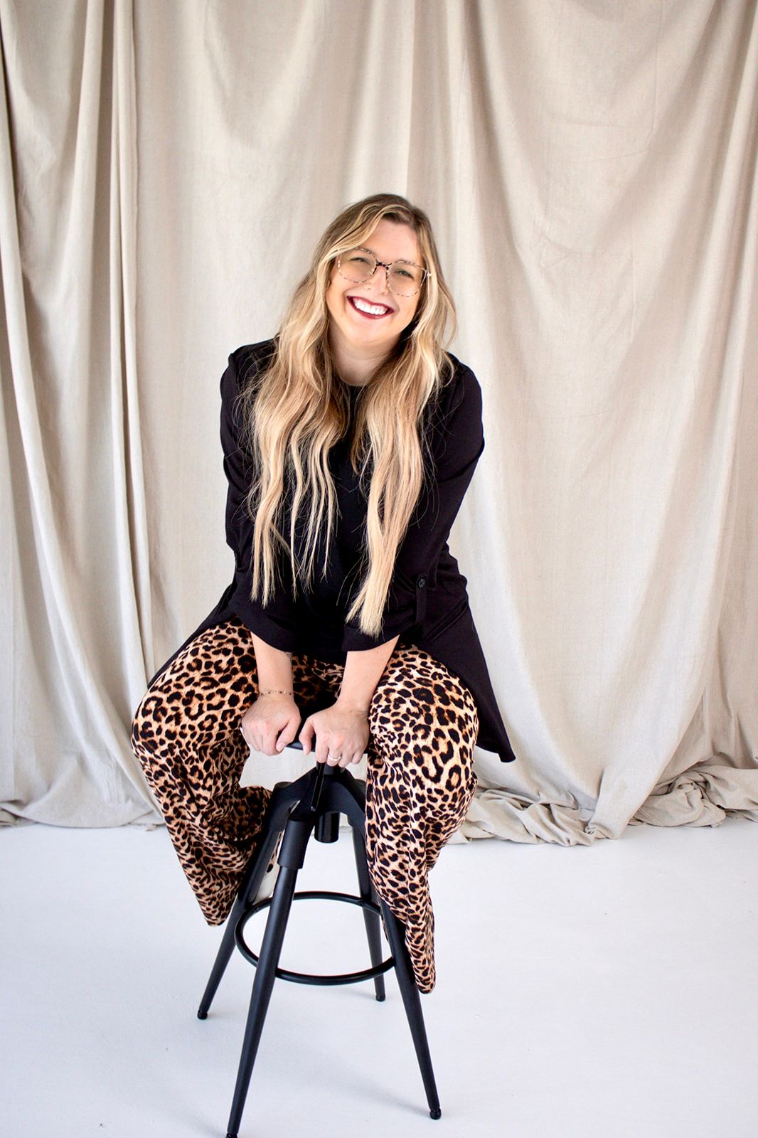 Smiling blonde woman in black blazer and leopard print pants sitting on a stool against a neutral draped backdrop.