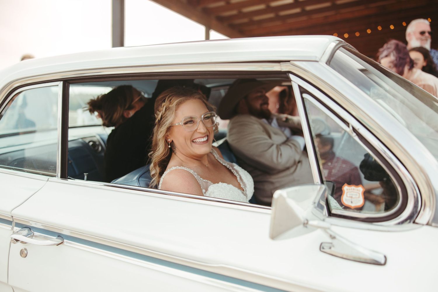 Smiling bride in white gown sits in a vintage white classic car on her wedding day.