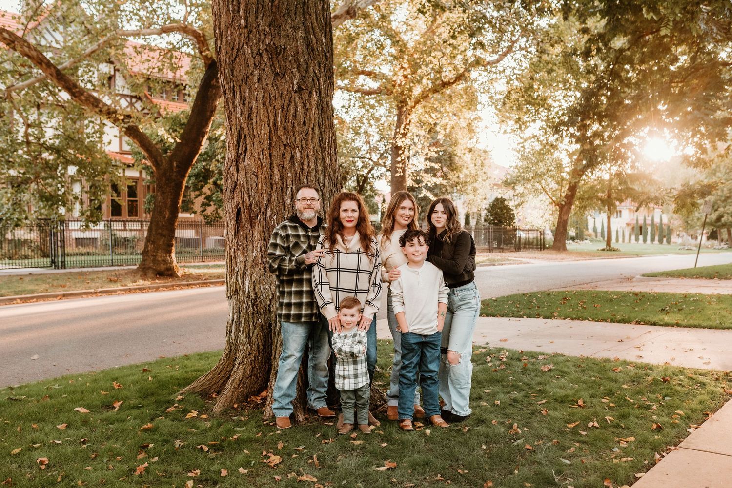 Family of six posing together under a large tree with golden fall foliage in a park.