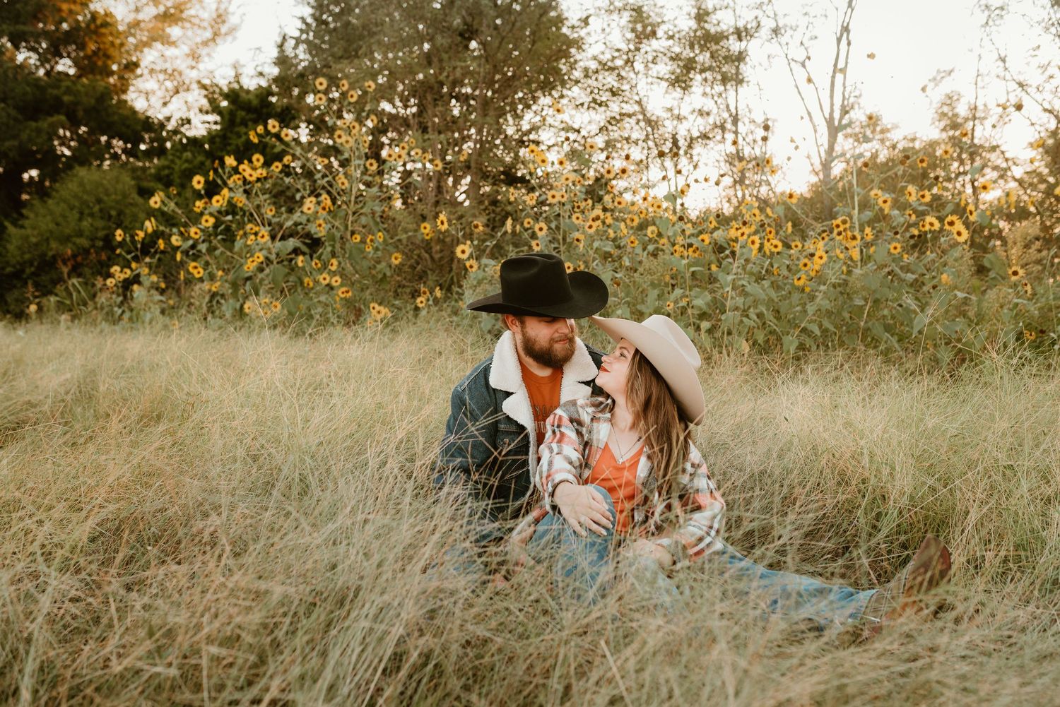 A couple in western hats sit together in tall grass surrounded by yellow wildflowers.
