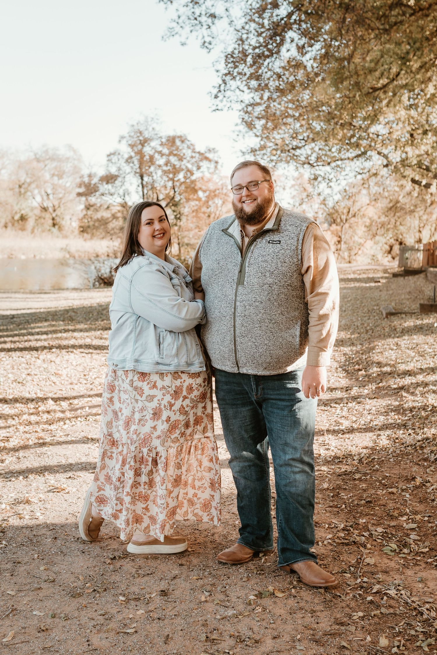 Smiling couple posing outdoors near a lake in autumn, woman in floral skirt and denim jacket.
