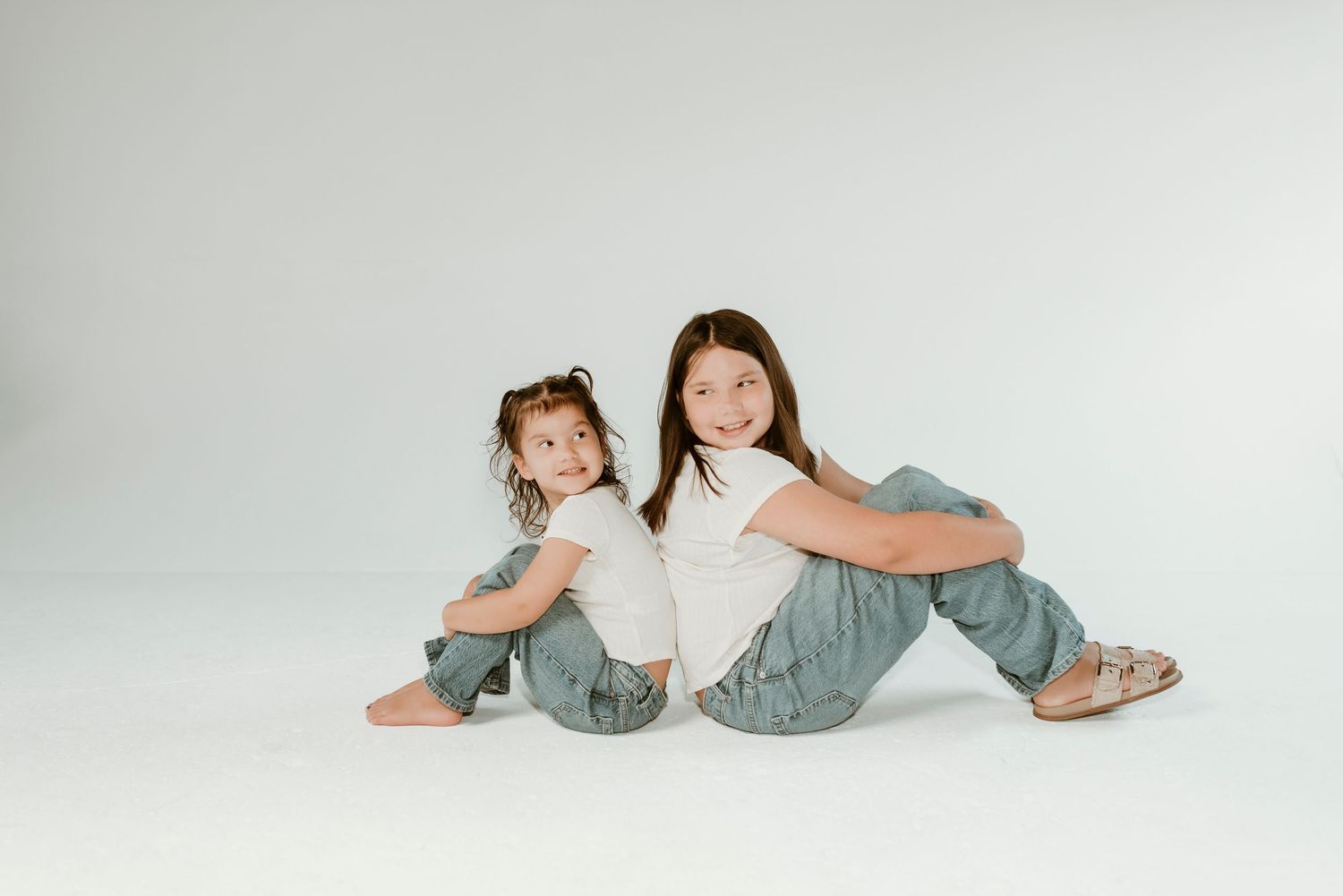 Two young sisters sitting back to back on white background wearing white tops and jeans.
