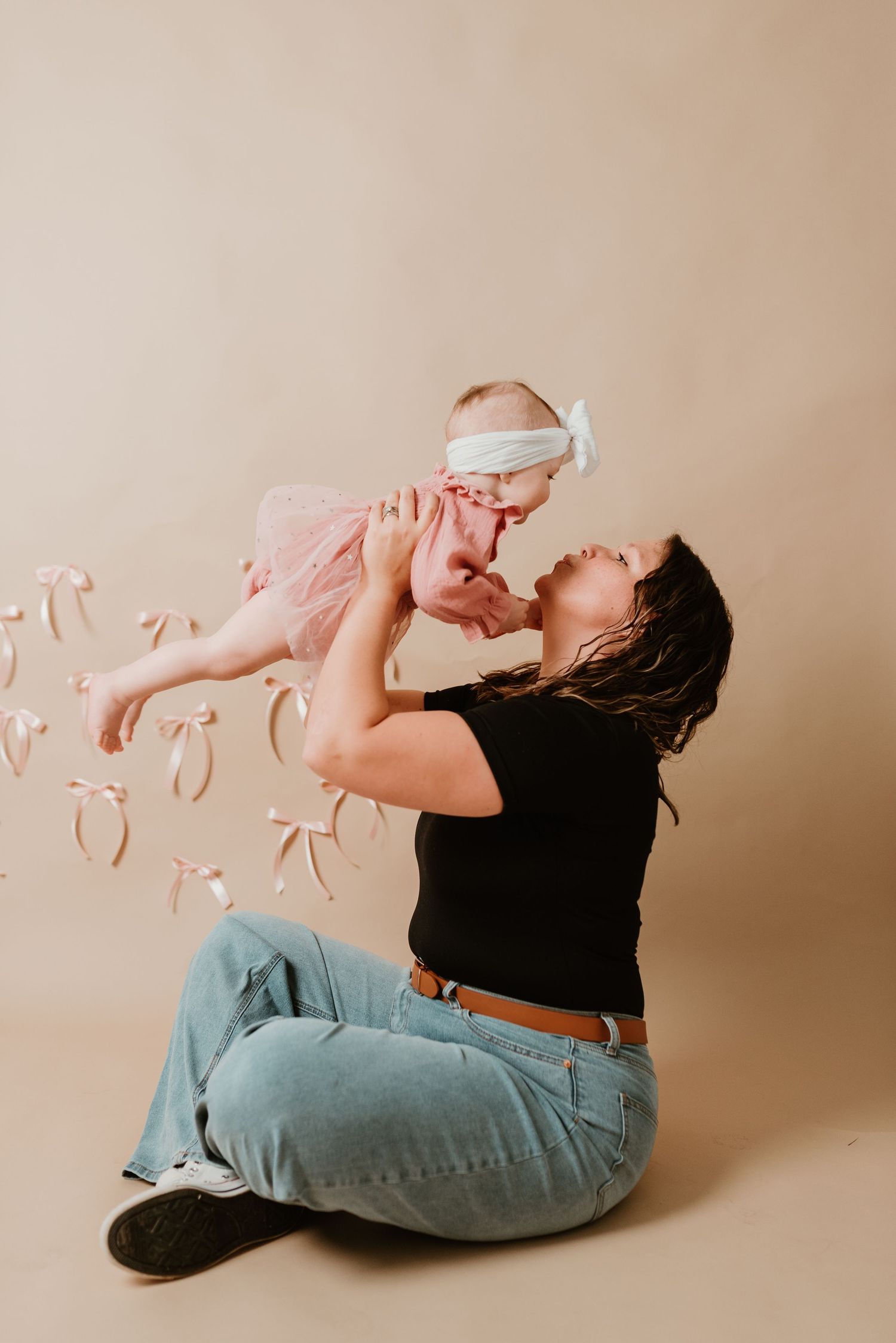 Mother lifts smiling baby girl in pink tutu overhead while sitting on floor with decorative pink bows floating around.
