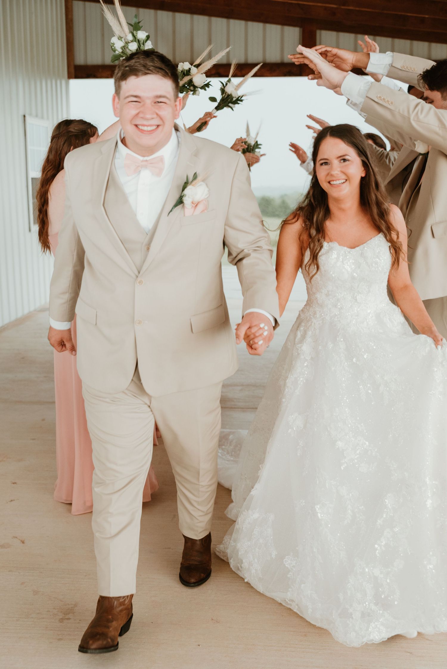 Smiling bride and groom in tan suit with pink bow tie walking through celebratory arch tunnel.