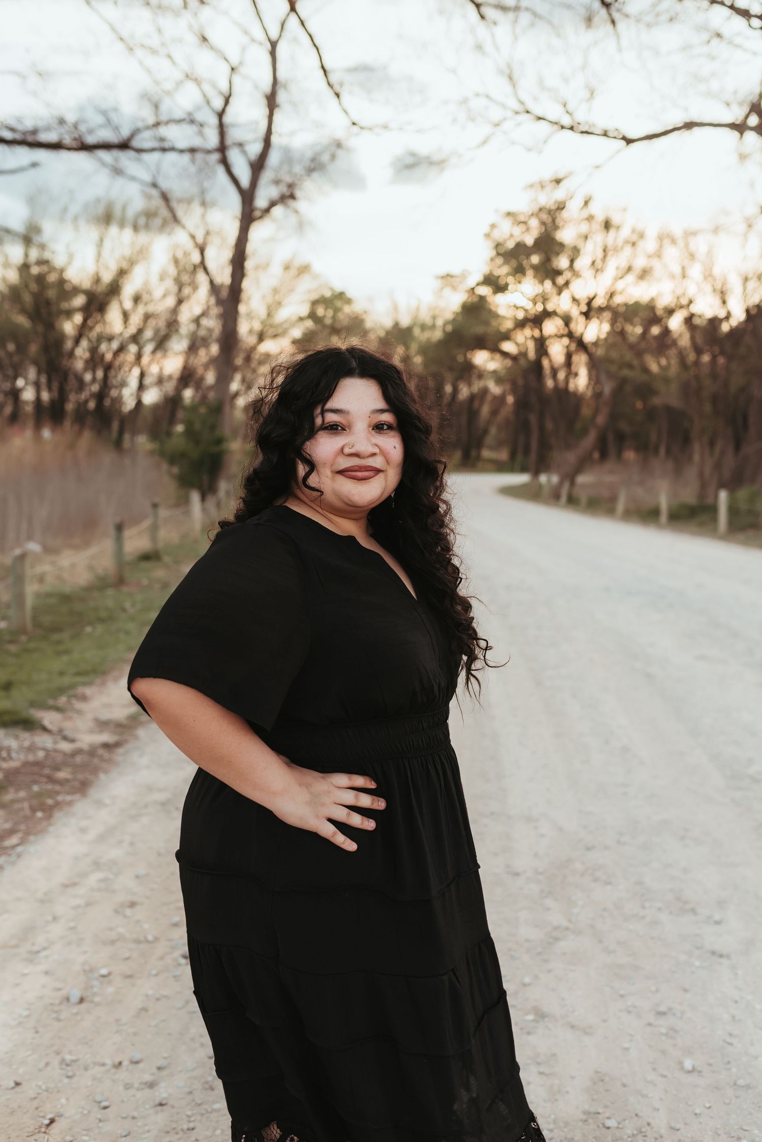 A woman in a black maxi dress poses confidently on a rural dirt road at sunset.