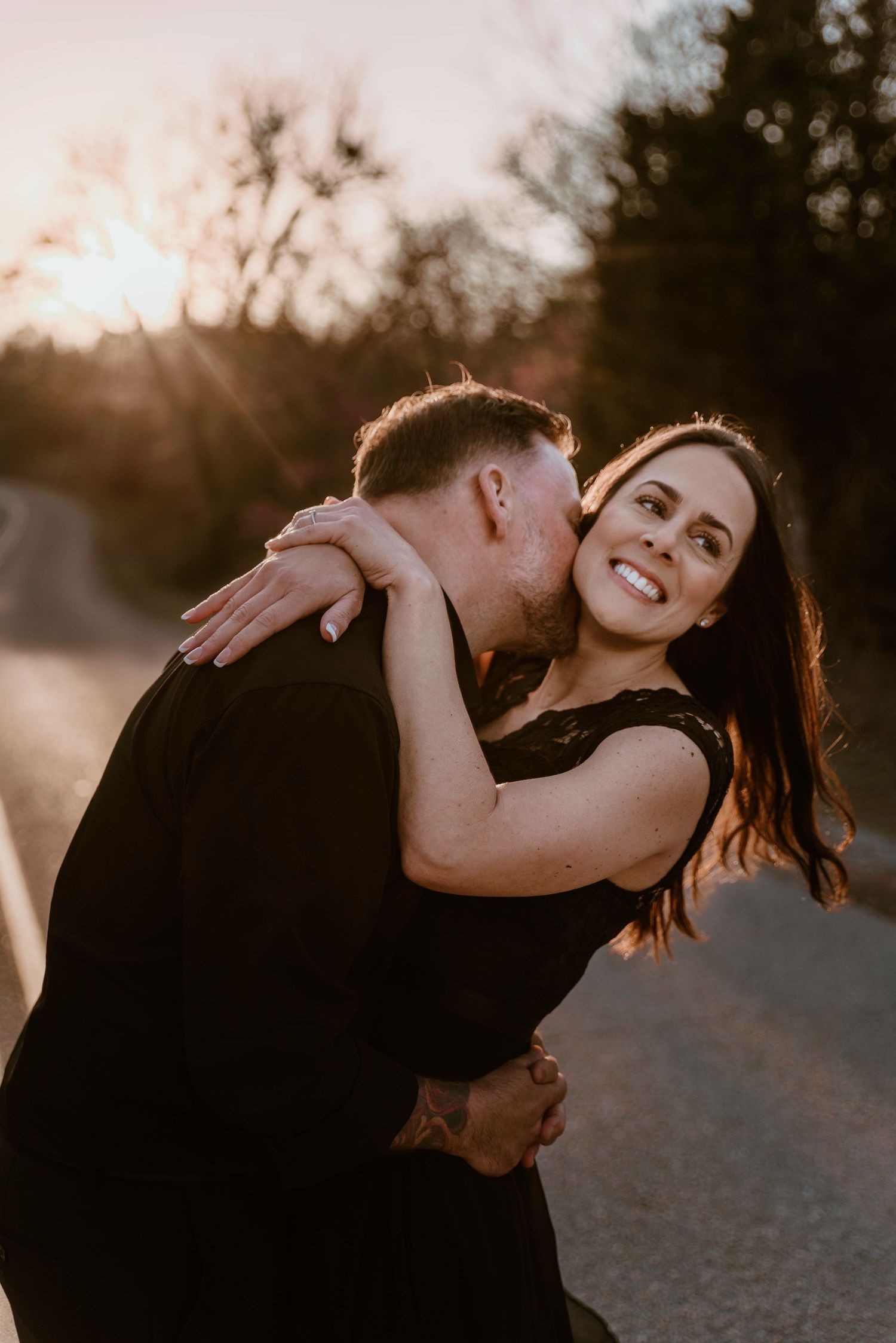 A man kisses a smiling dark-haired woman on the cheek during a glowing sunset roadside photo session.