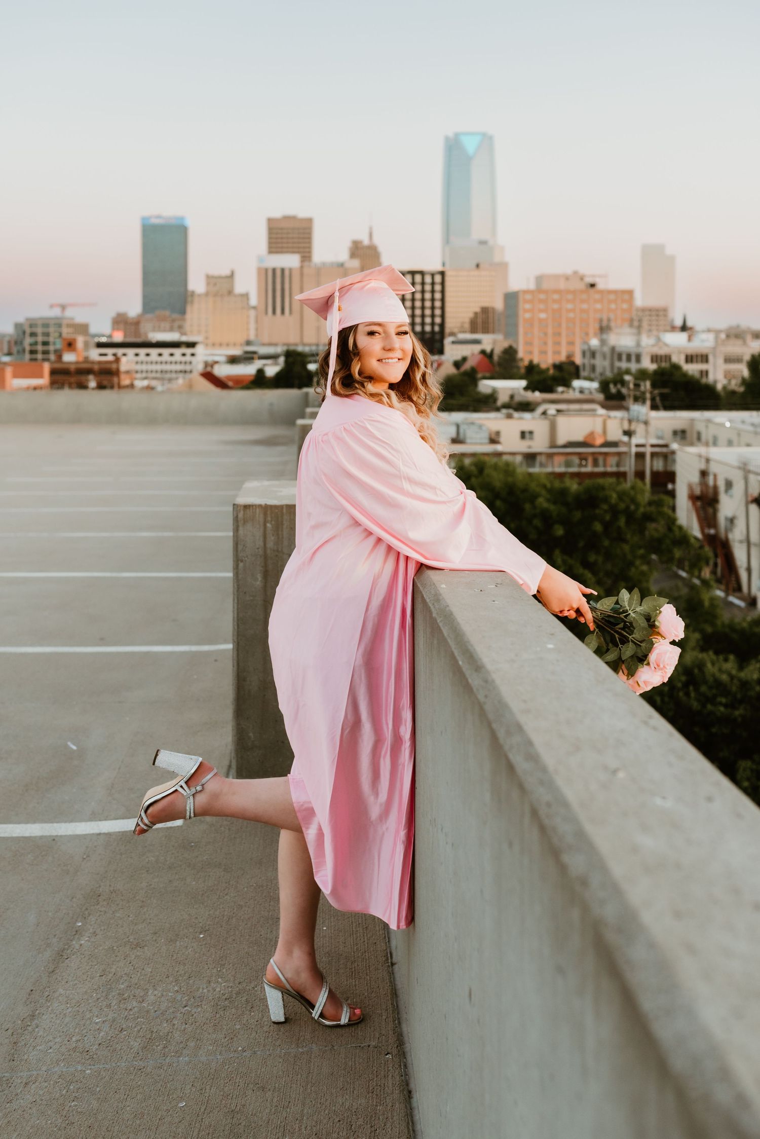 Graduate in pink cap and gown posing on rooftop ledge with city skyline at dusk.
