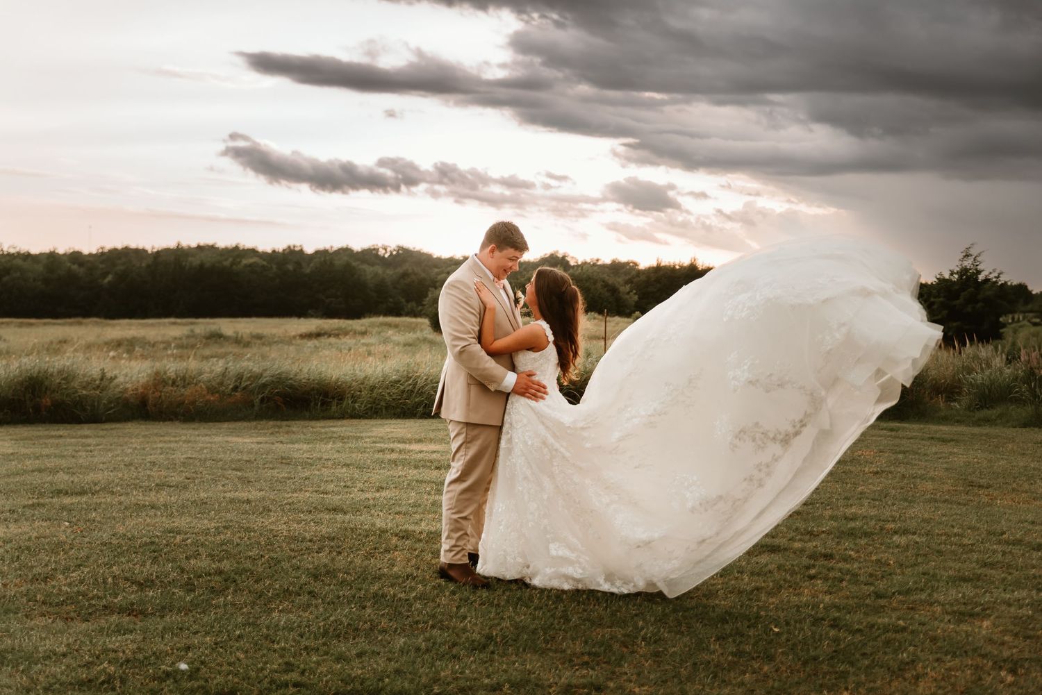 Bride and groom share romantic embrace outdoors at sunset with dramatic cloudy sky and flowing wedding dress.