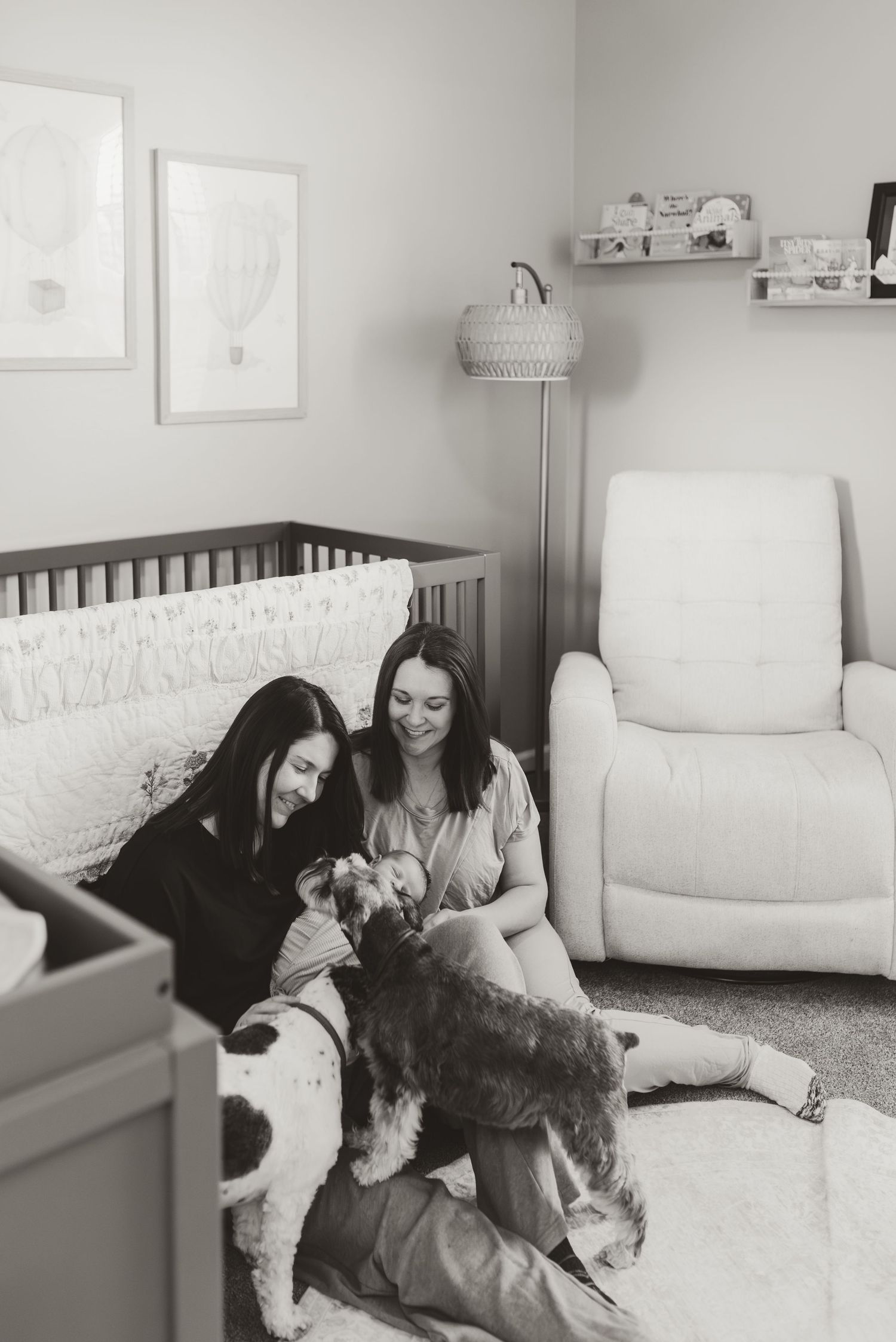 Two women sit on a nursery floor smiling and petting their dogs in a cozy black and white photo.