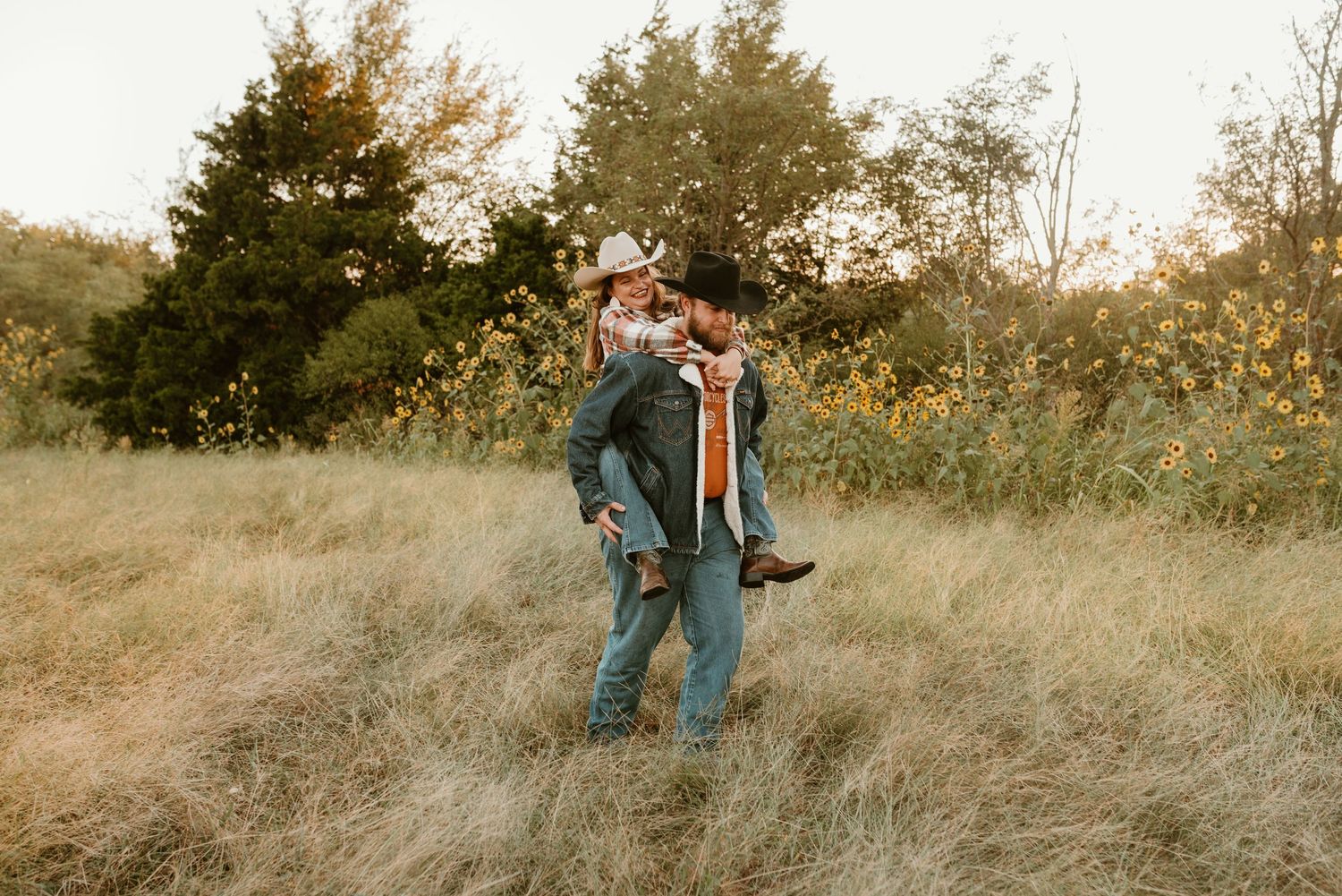 A man gives his partner a piggyback ride through a sunny meadow with yellow wildflowers.