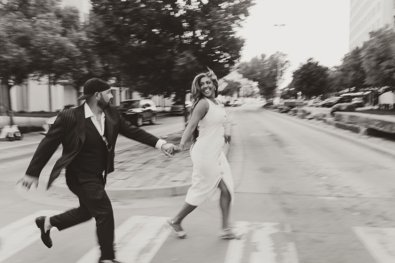 Black and white photo of a joyful couple holding hands and running across a city street crosswalk.